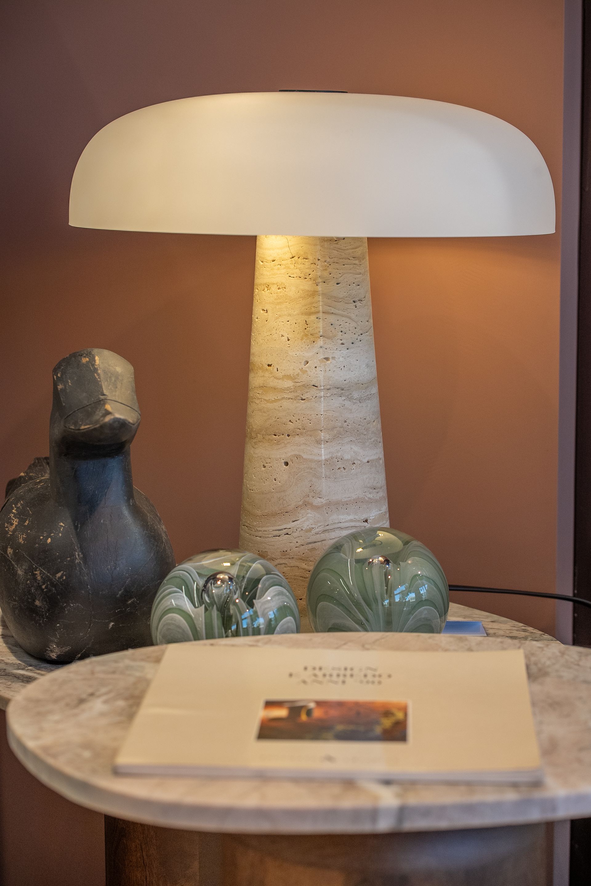 Lampada da tavolo con paralume bianco su un tavolo rotondo di marmo, con oggetti decorativi e un libro immersi in una luce calda.