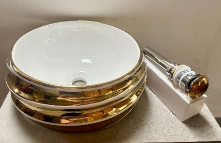 Lavabo de baño de cerámica blanca y dorada con tapón de drenaje dorado sobre una superficie gris.