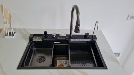 Fregadero de cocina negro con accesorios integrados, escurridor de acero inoxidable y grifo alto sobre encimera blanca.