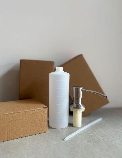 Dispensador de jabón blanco, botella y tubo junto a cajas de cartón sobre una superficie clara.