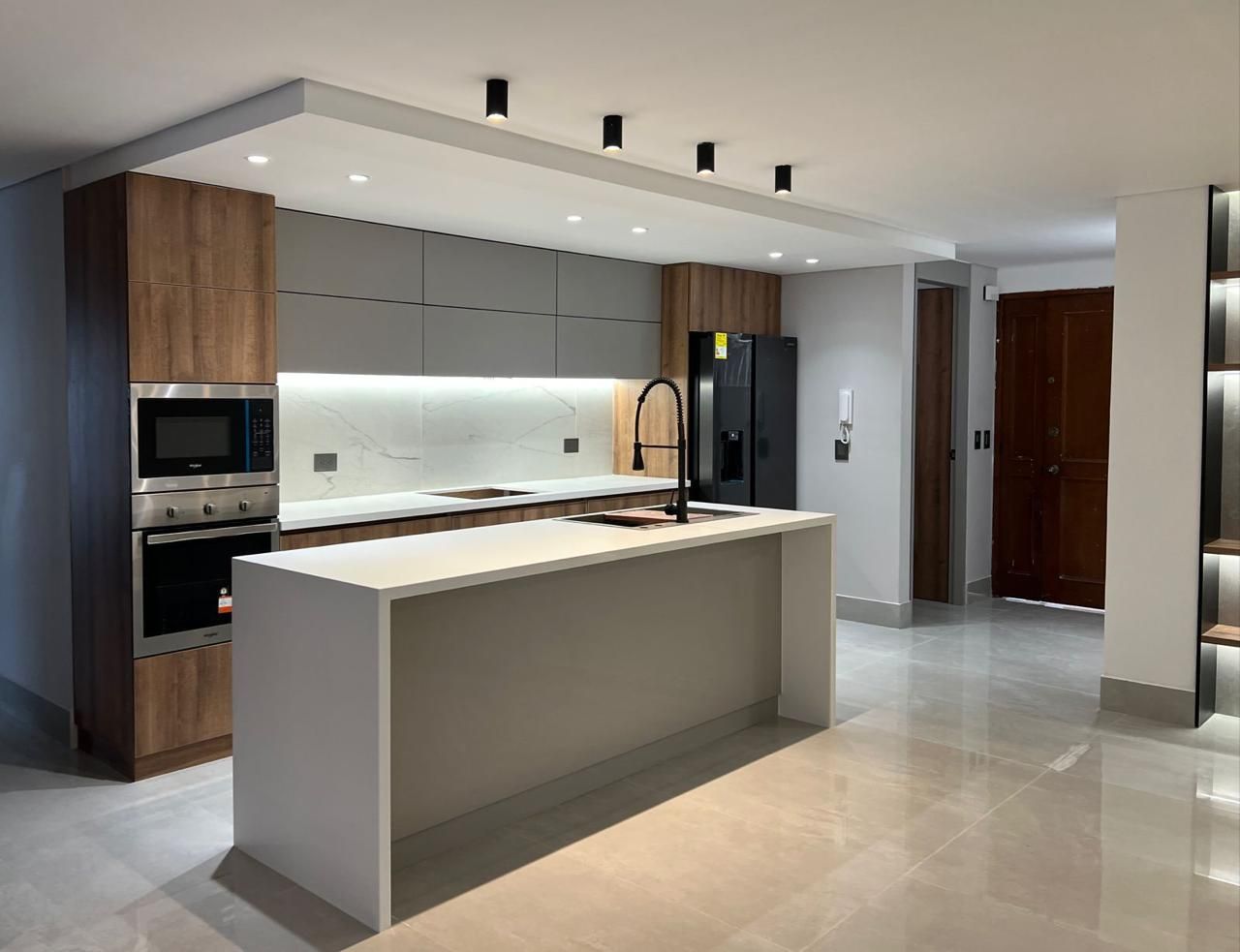 Cocina moderna con isla blanca, gabinetes grises, detalles de madera y electrodomésticos negros.