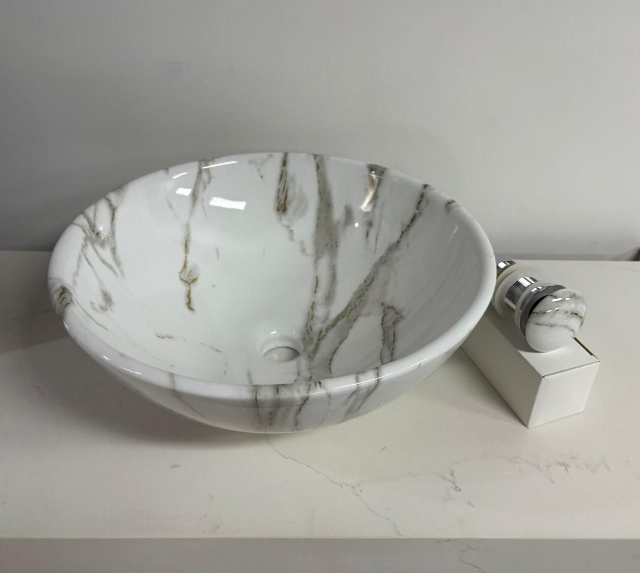 Lavabo con diseño de mármol blanco y tapón de drenaje plateado, sobre una encimera blanca.
