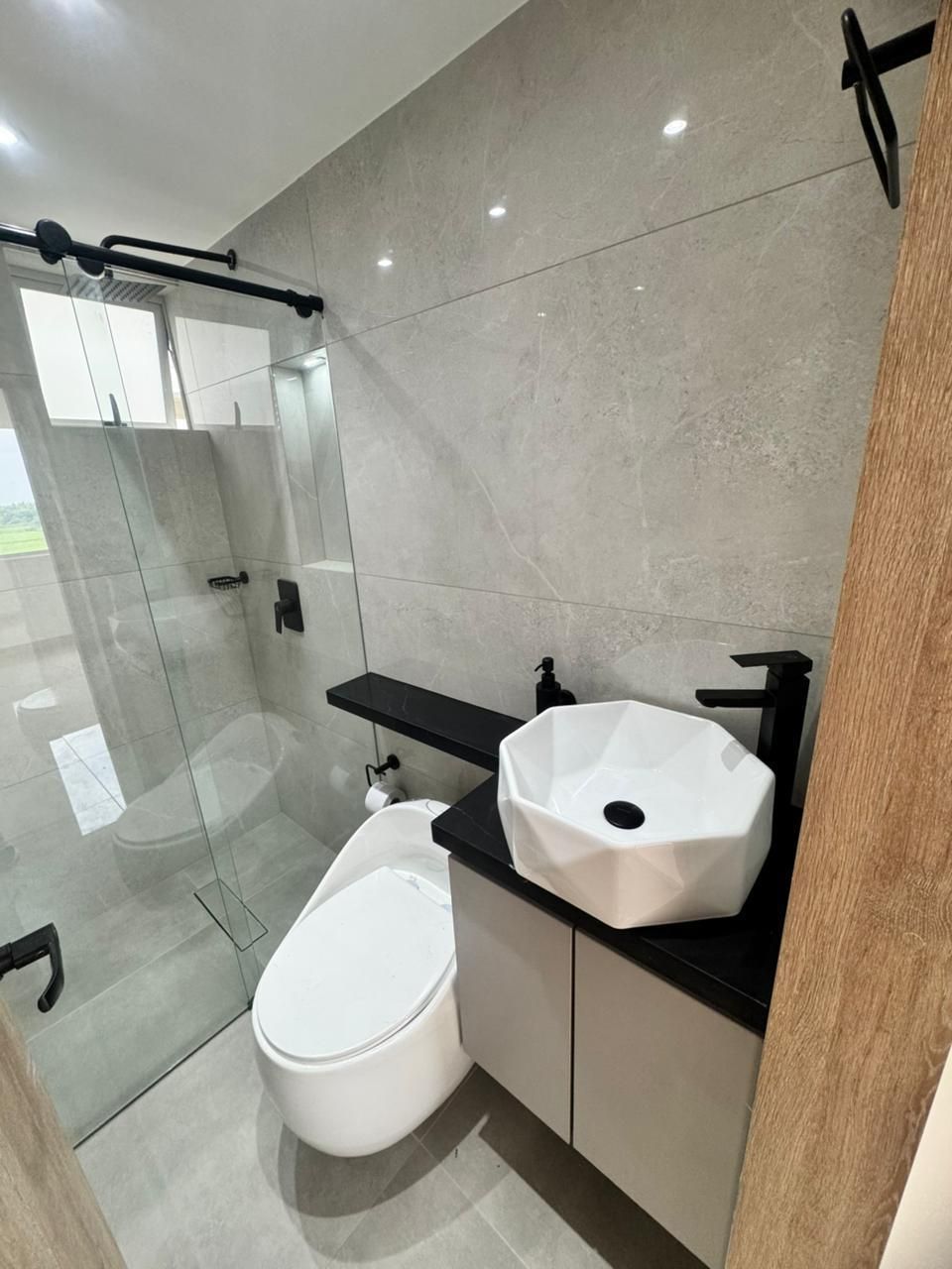 Baño moderno con azulejos grises, accesorios negros, lavabo geométrico y ducha de cristal.