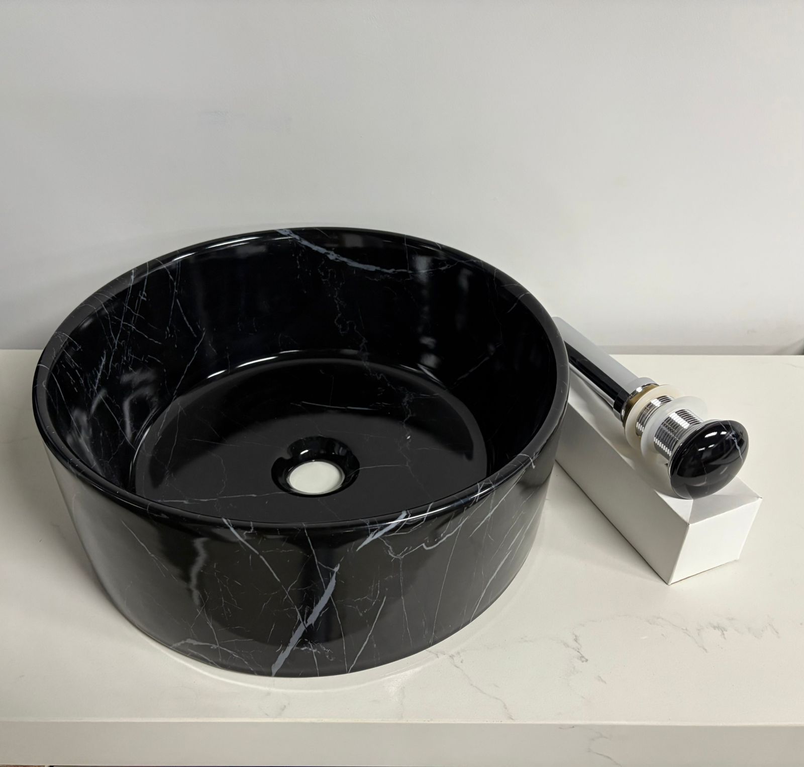 Lavabo de baño redondo de mármol negro y desagüe cromado, colocado sobre una superficie blanca.