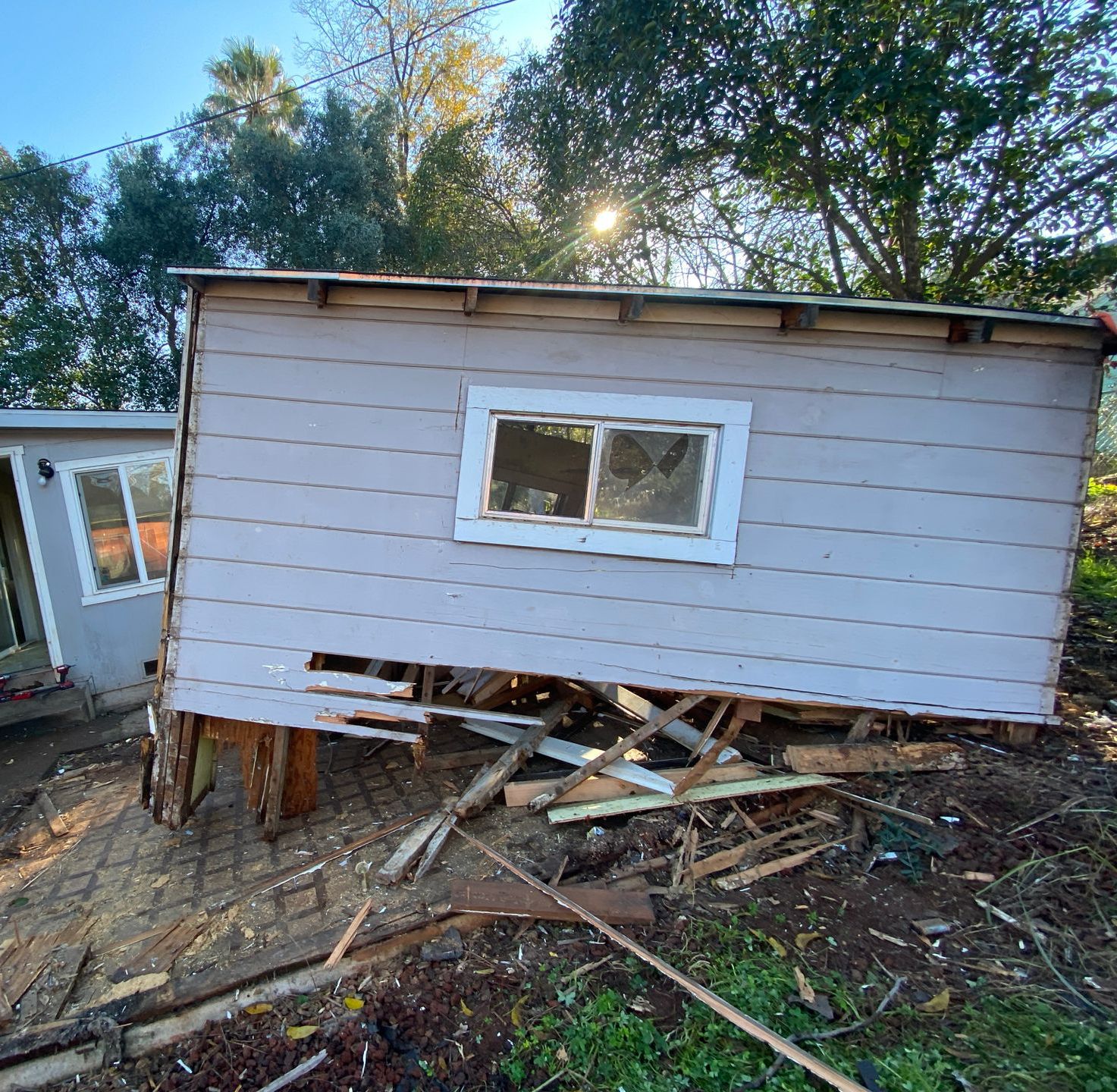 Tilted gray shed on a sloped yard, with a broken lower support and trees behind it.