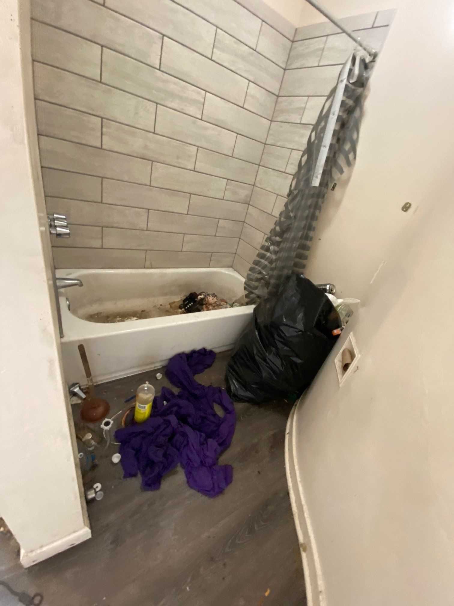 Cluttered bathroom with a bathtub, black trash bags, purple cloth, and cleaning supplies on the floor.