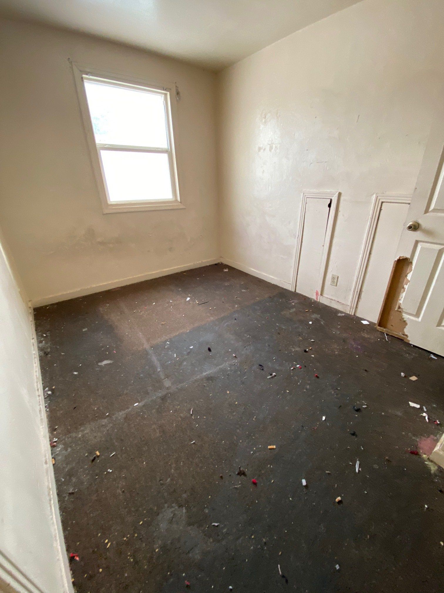 Empty room with beige walls, a window, and debris scattered on a dark floor.