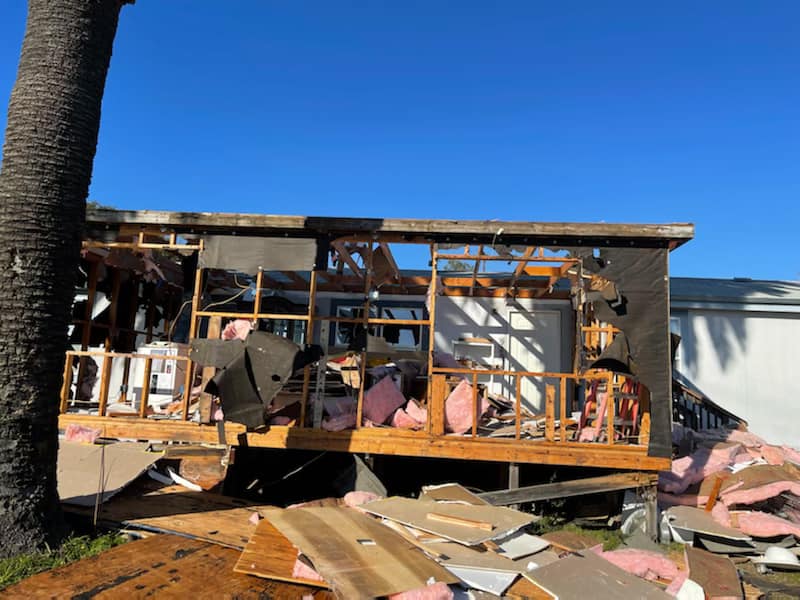 Damaged house with blown-out walls and debris scattered in front under a clear blue sky