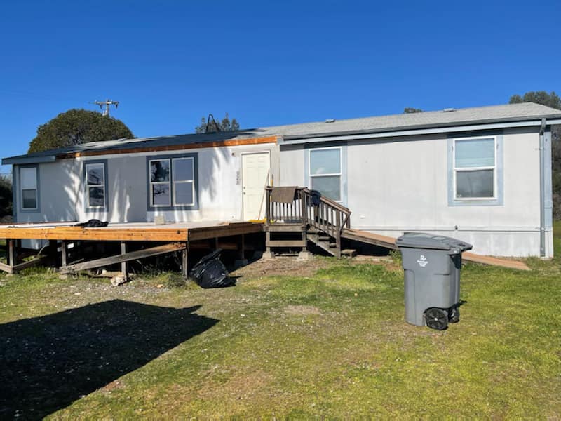 Single-story white mobile home with a small wooden deck on a grassy lot under a clear blue sky.