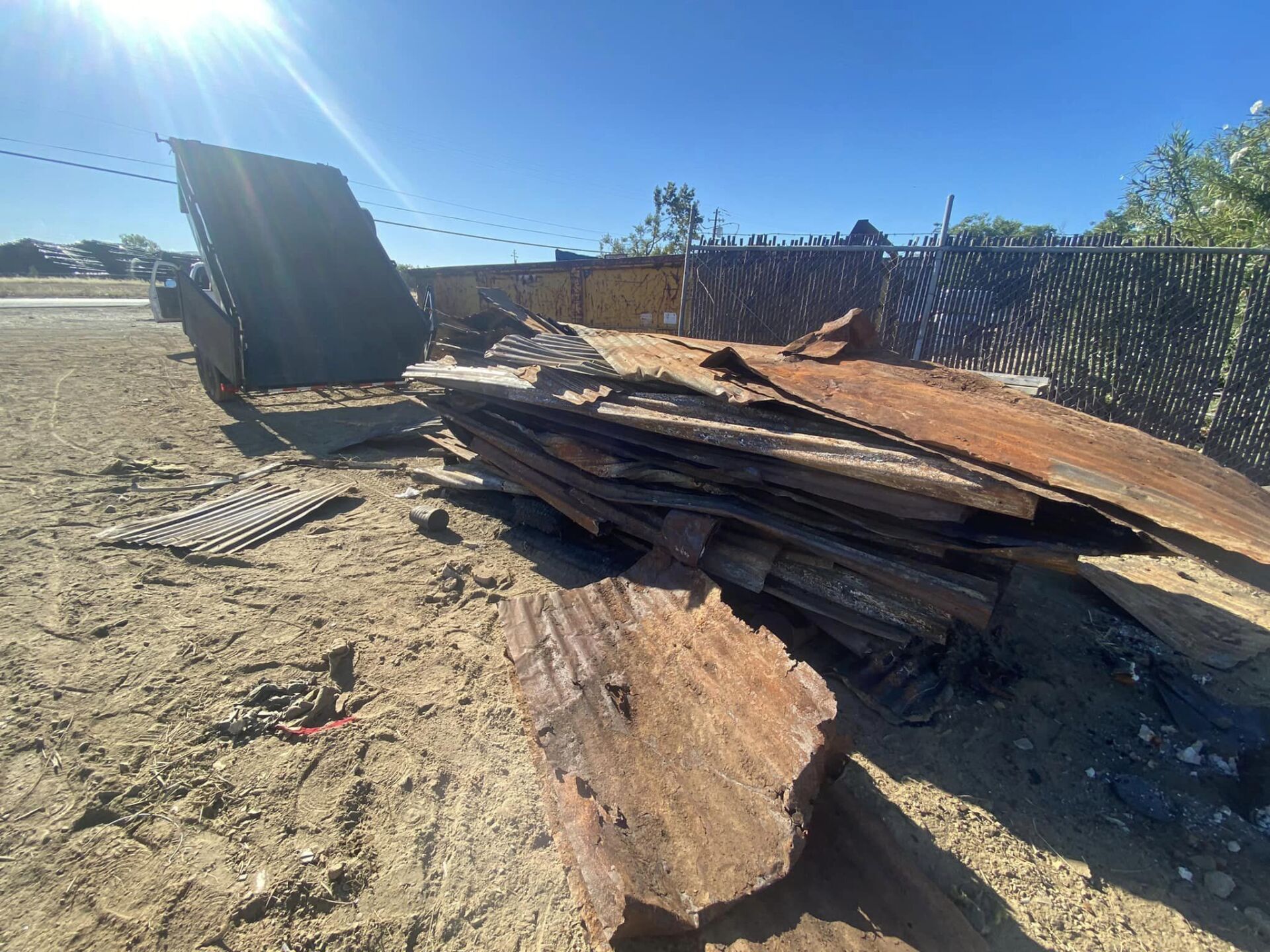 Rusty corrugated metal debris piled on sandy ground beside a chain-link fence under a blue sky
