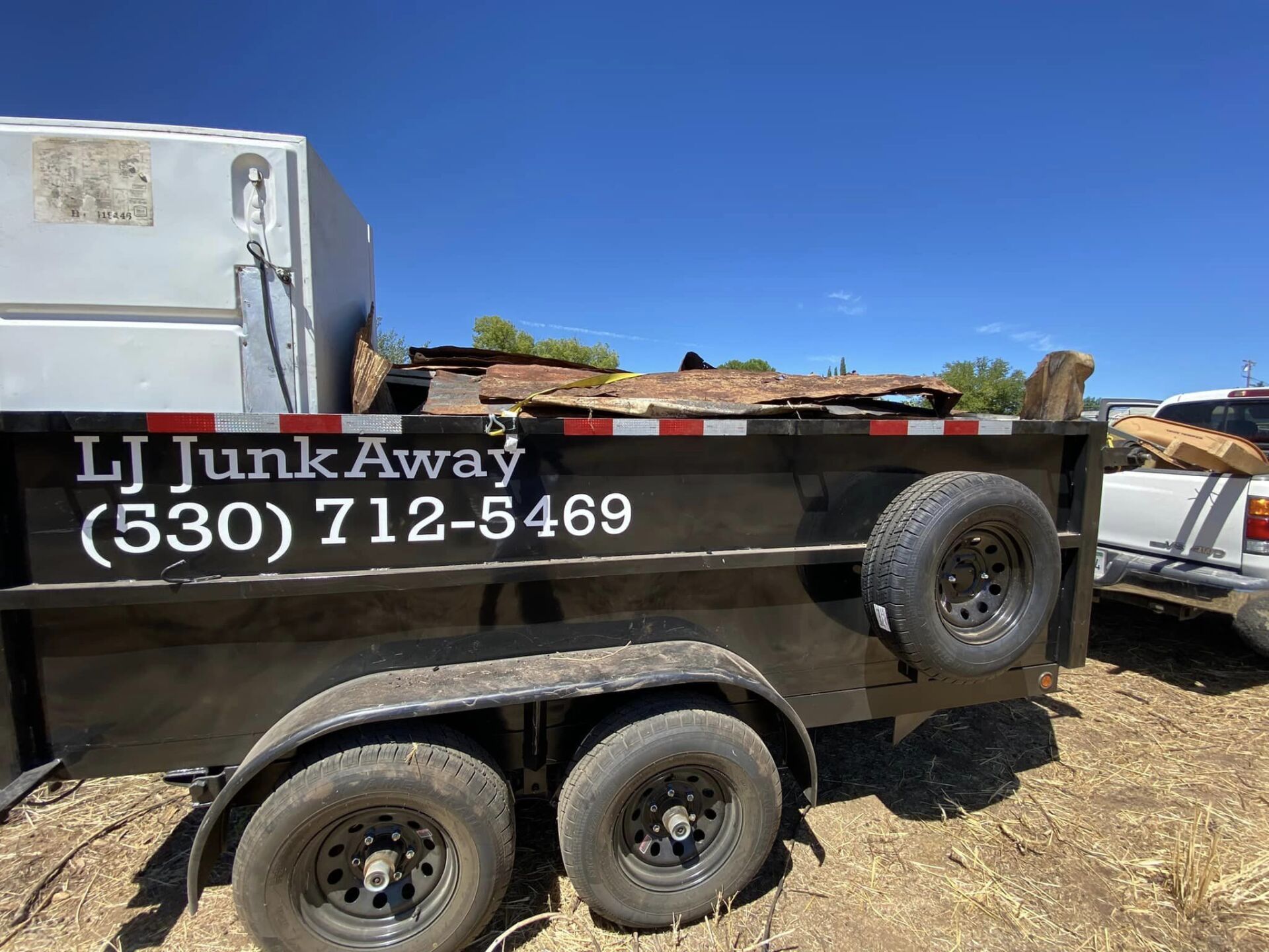 Black junk removal trailer with “I Junk Away” and phone number parked on dry ground under blue sky.
