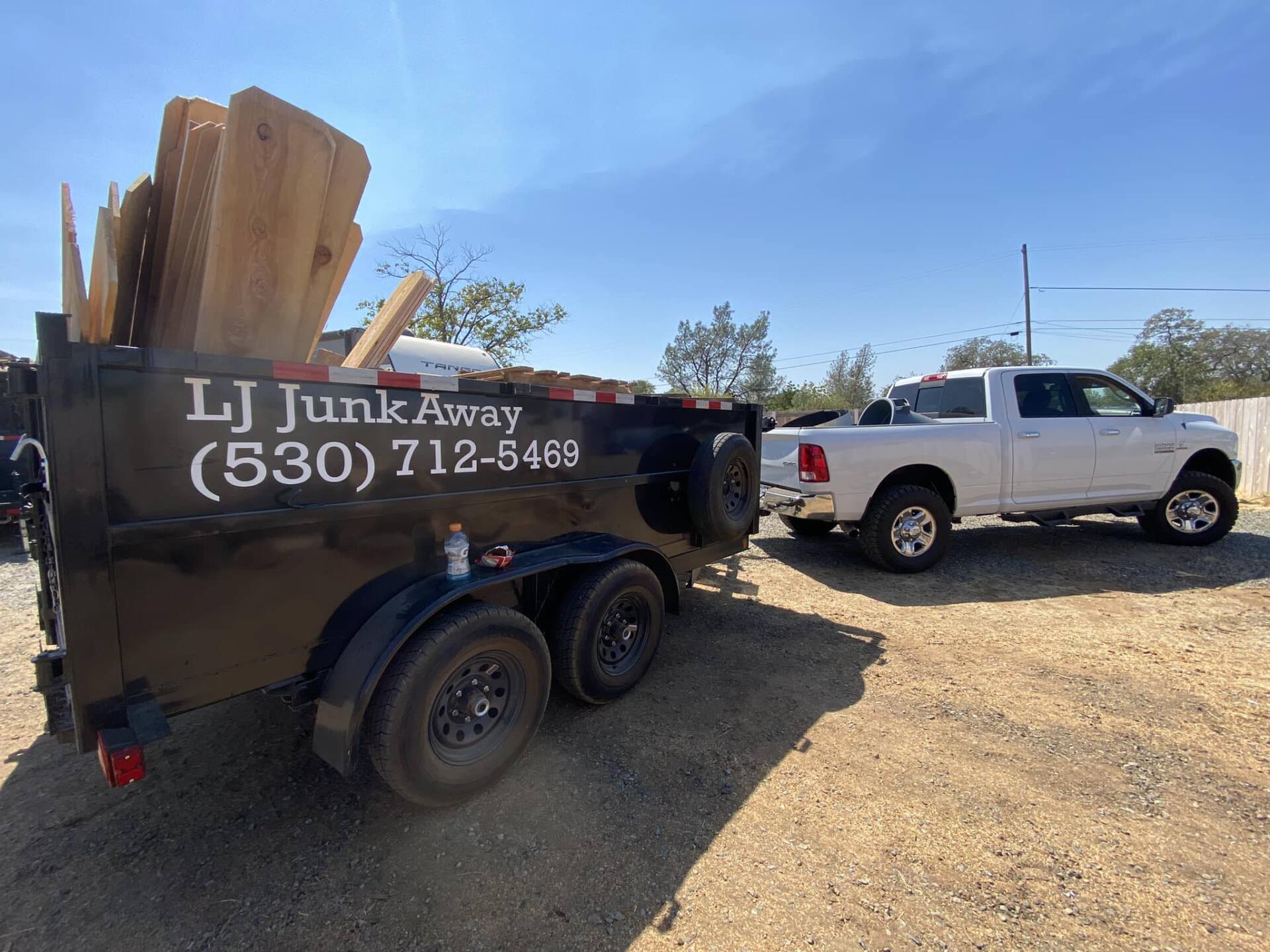 Dump trailer with lumber and white pickup truck on a gravel lot under a blue sky