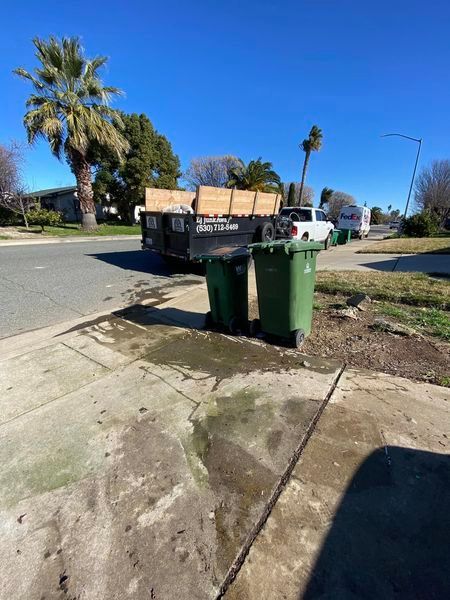 Two green trash bins on a sidewalk beside a suburban street under a clear blue sky.