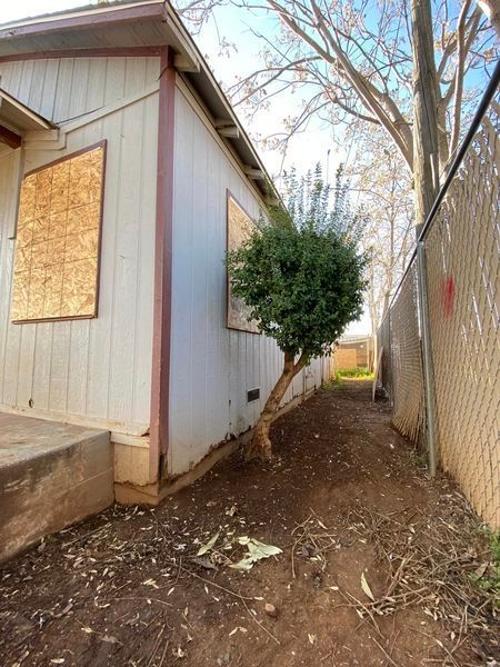 Narrow dirt path beside a boarded-up white building and chain-link fence, with a bush and bare tree branches.