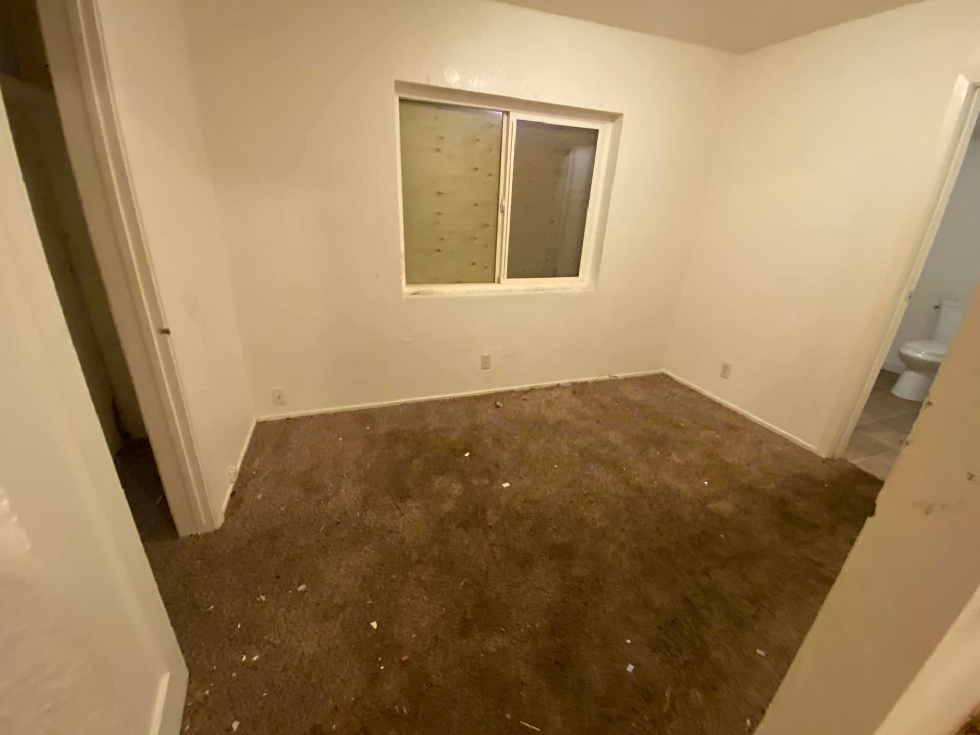 Empty small room with brown carpet, white walls, a window, and a bathroom doorway on the right