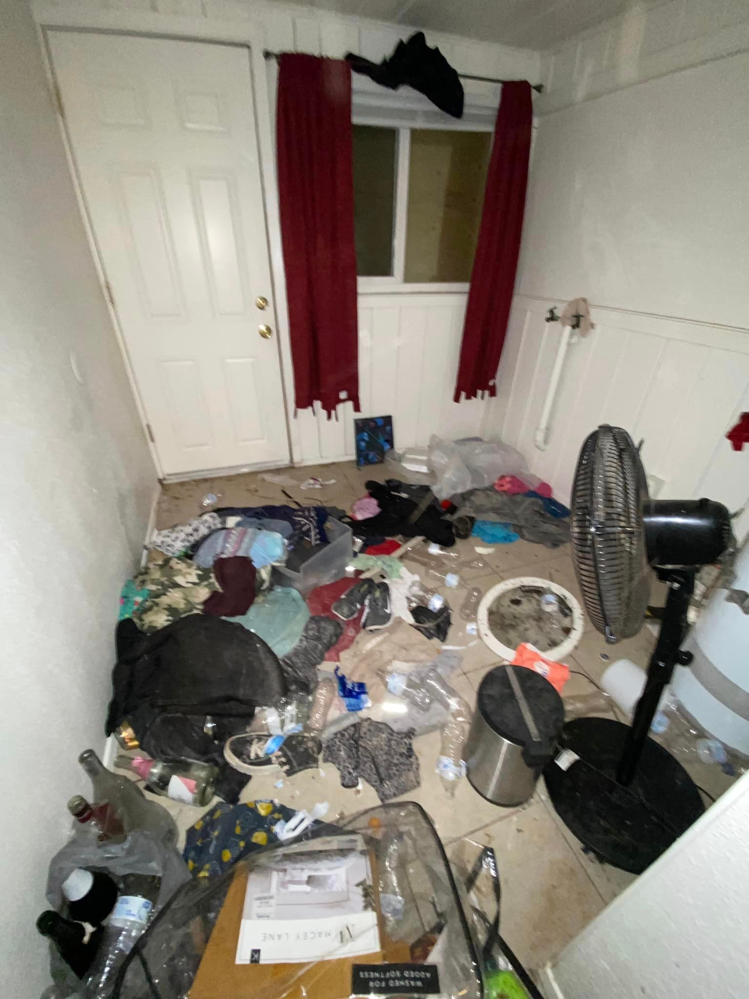 Cluttered, messy room with trash and scattered belongings on the floor, window with red curtains, and a fan