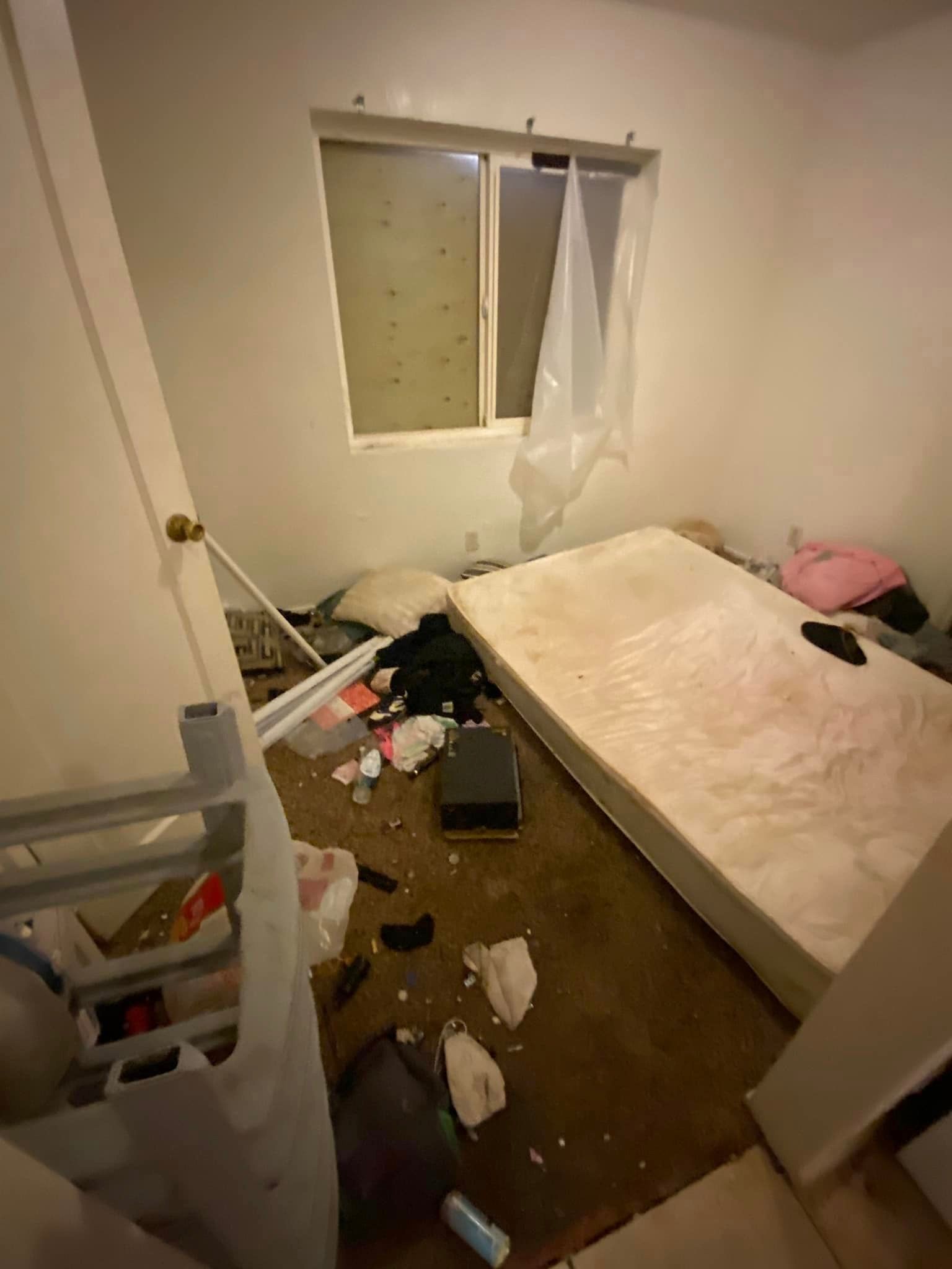 Messy bedroom with unmade bed, clothes and trash scattered on the floor, and a partly open window curtain