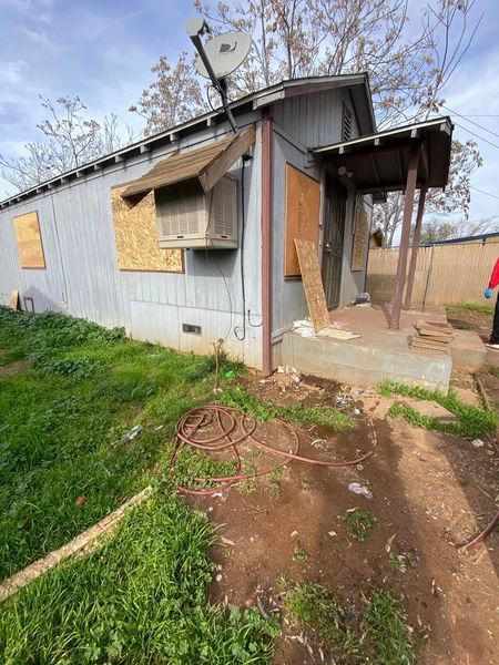 Small boarded-up gray house with a damaged porch, broken windows, and a dirt path in front.