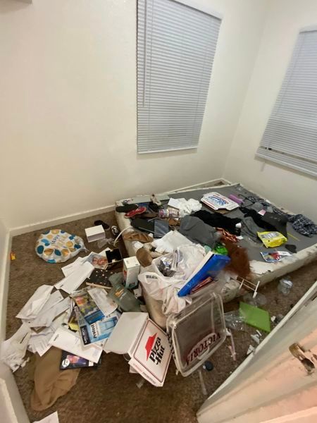 Cluttered room with papers, boxes, and trash scattered across the floor near two windows with blinds.