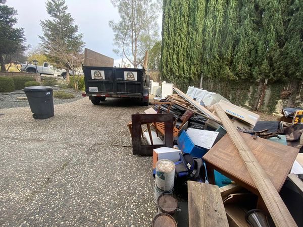 Backyard gravel area with a black dumpster, bins, and a pile of discarded wood and debris