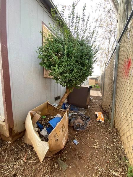 Trash piled in a narrow alley beside buildings and a chain-link fence.