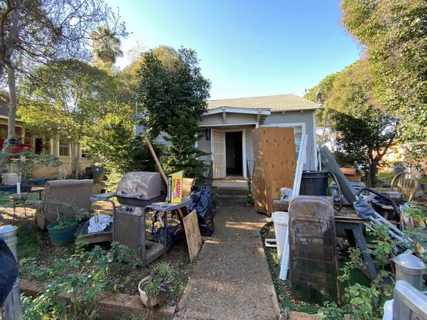Front yard with scattered furniture and debris, boarded house entrance, and a ladder leaning on the right side