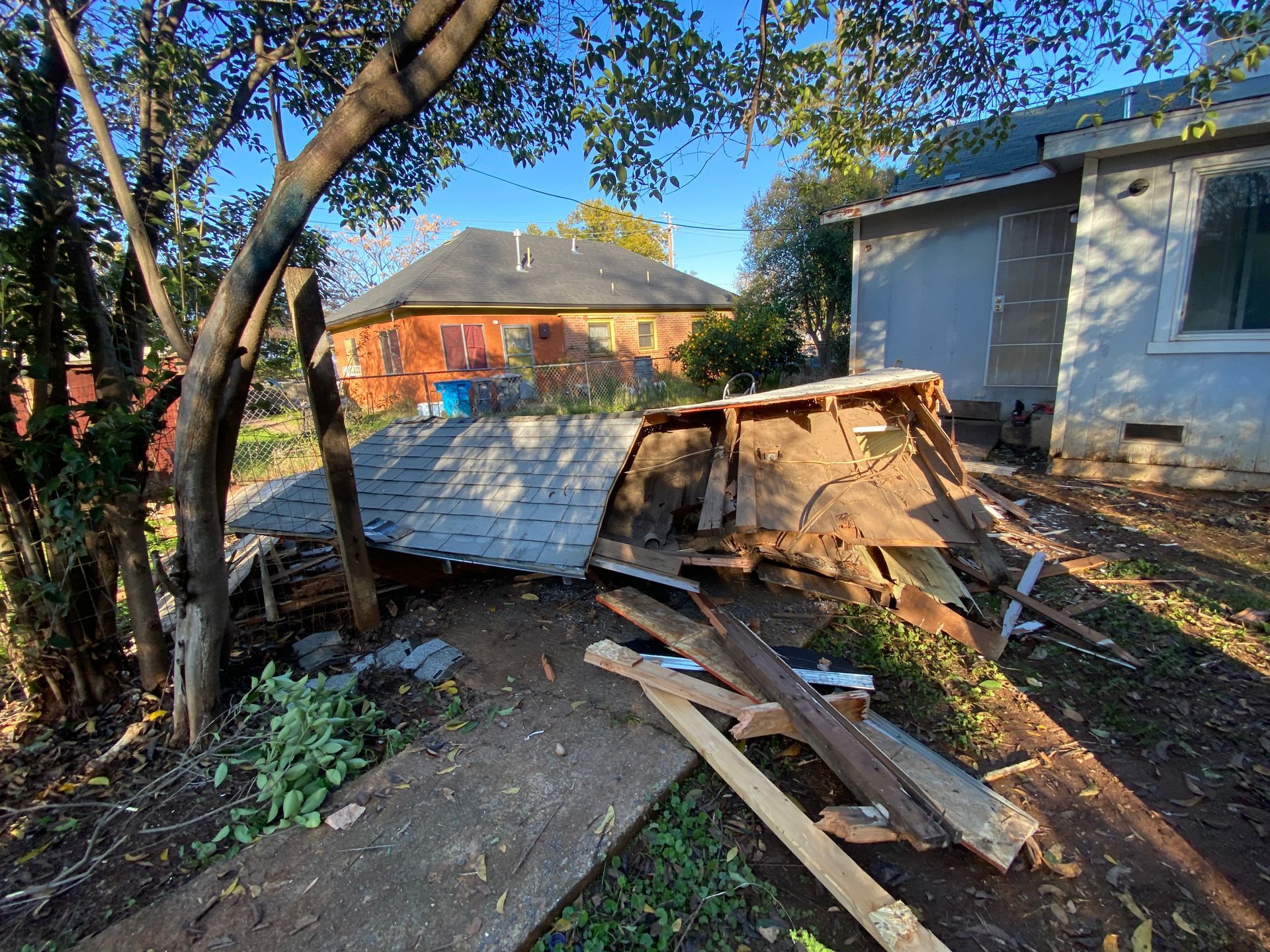 Collapsed wooden shed in a backyard beside a house, with scattered debris and trees overhead.