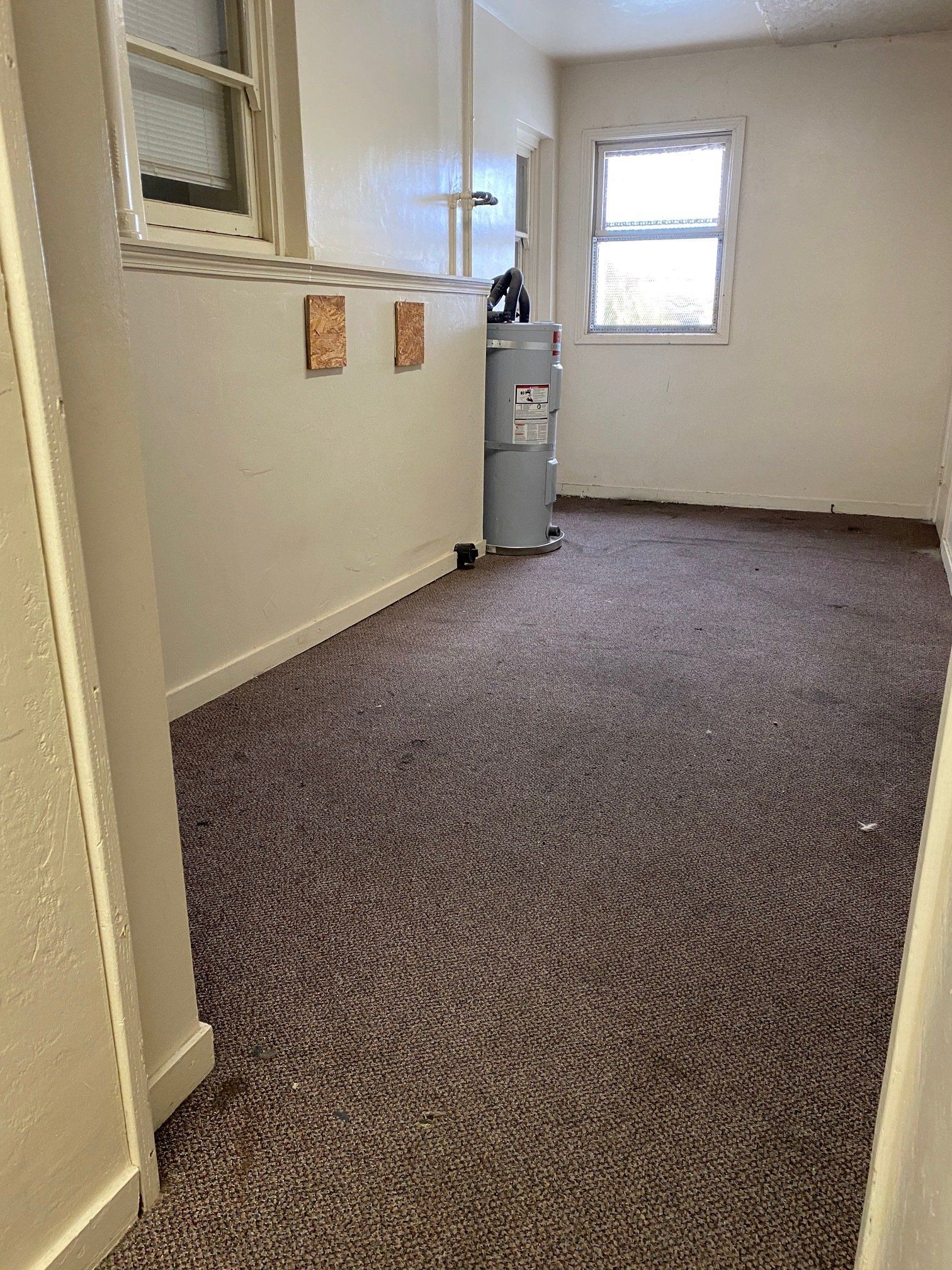 Empty carpeted room with beige walls, two windows, and a water heater near the back wall