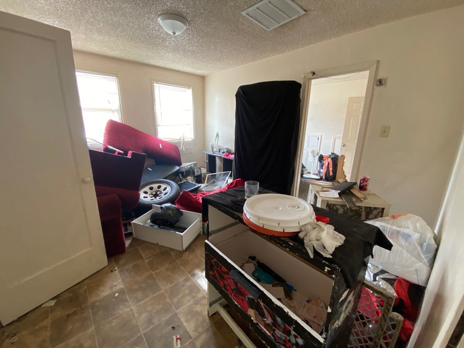 Cluttered room with scattered furniture, boxes, and household items near windows and an open door.