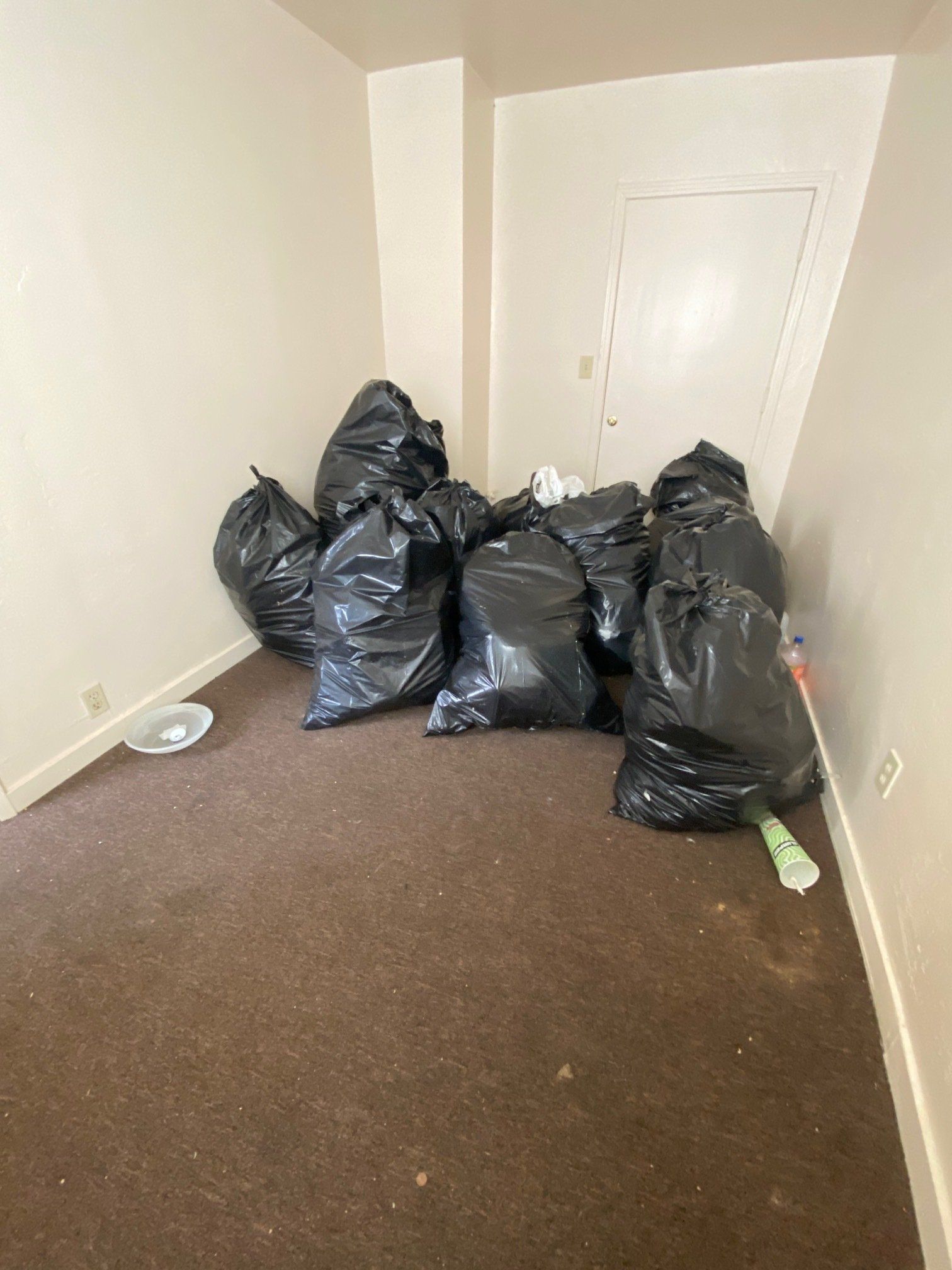 Small empty room with several black trash bags piled in the corner on brown carpet.