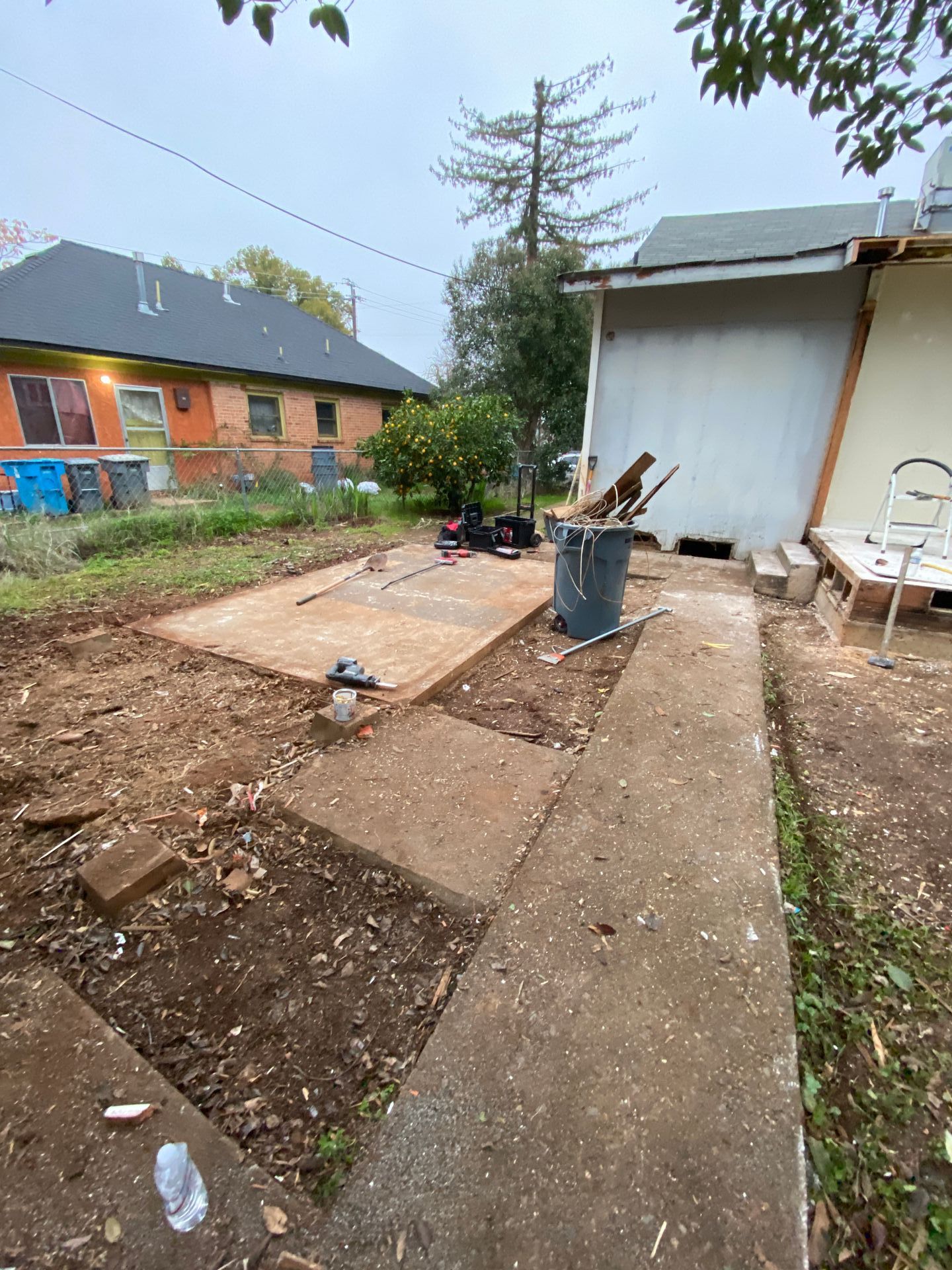 Backyard patio with concrete slabs, dirt patches, a bucket of tools, and a shed beside a house