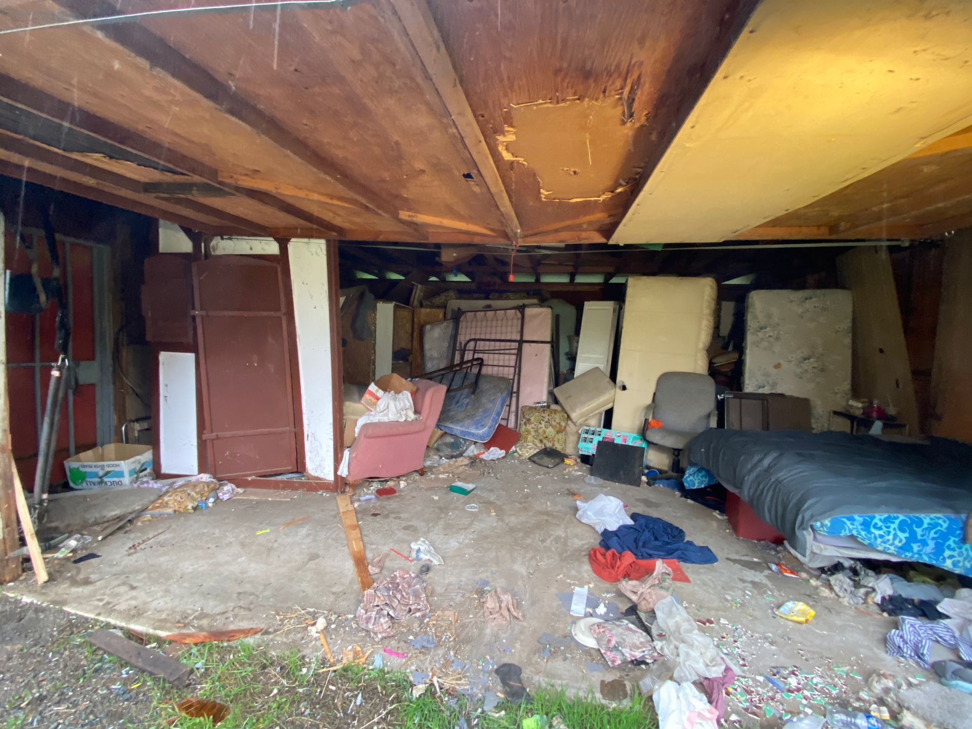 Cluttered basement with exposed beams, scattered debris, furniture, and plastic-covered items under a low ceiling