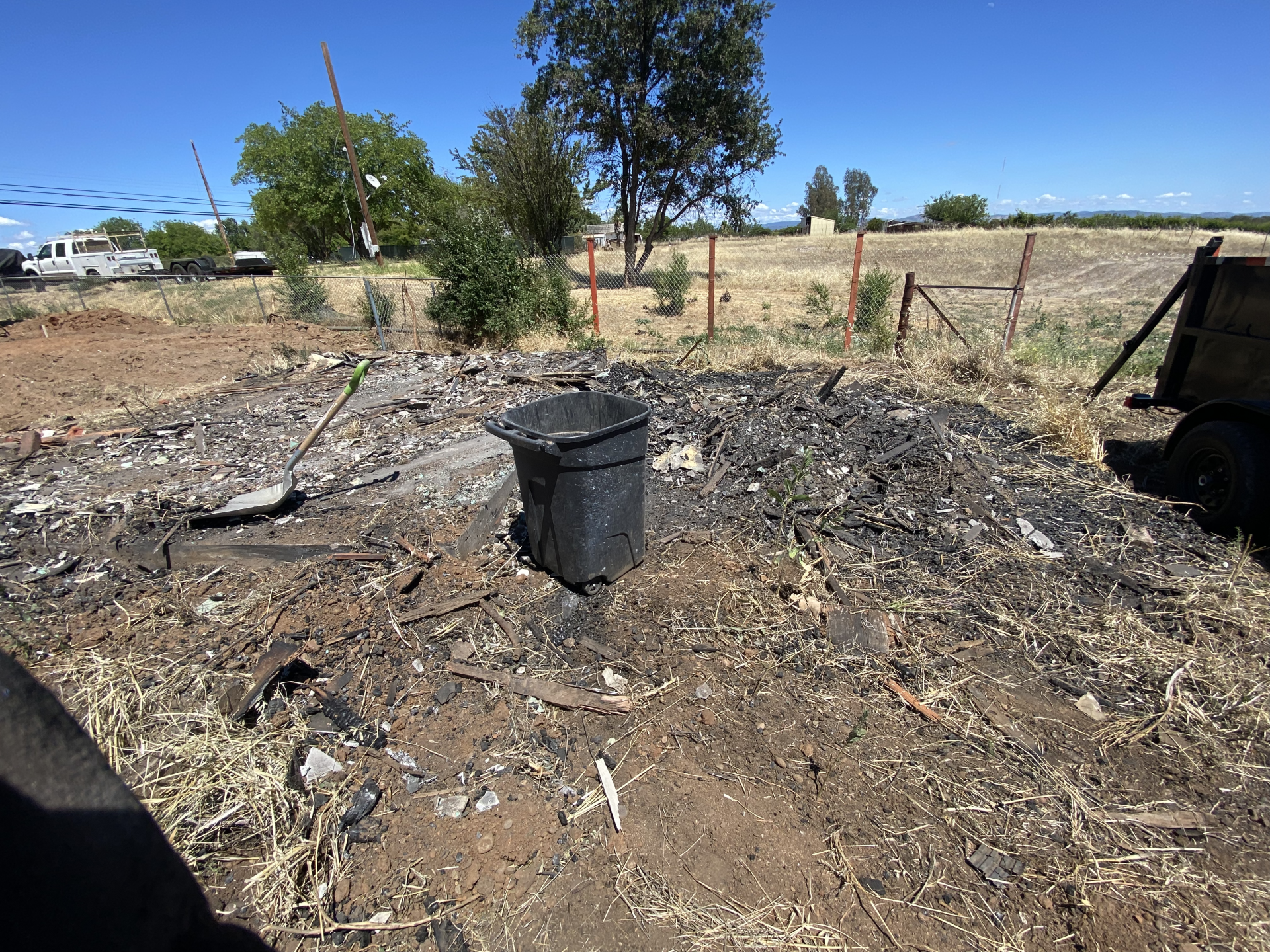 Burned debris pile with a black barrel in a dry rural yard under a clear blue sky