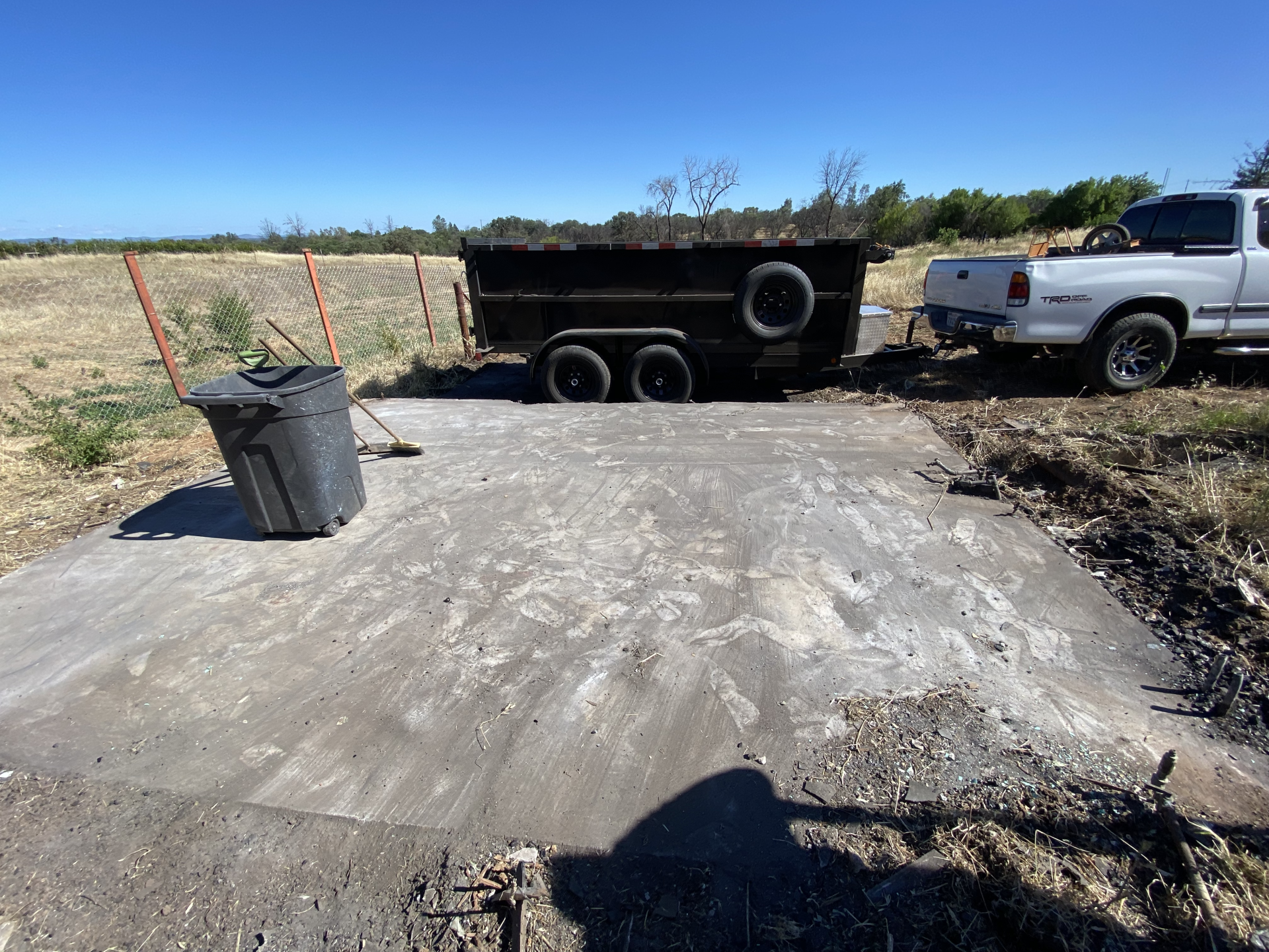 Concrete driveway with a black trailer, white pickup truck, and trash bin under a clear blue sky