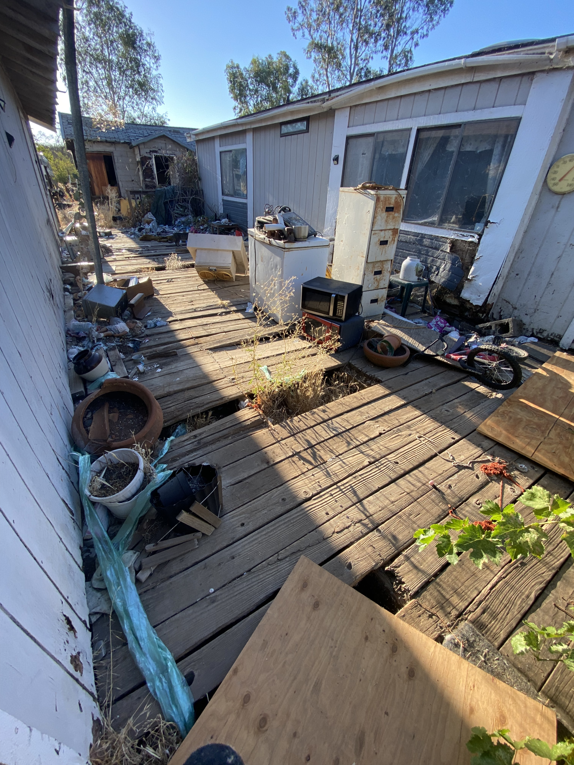 Cluttered wooden deck beside a house, with tools, debris, and a white cabinet in bright sunlight.