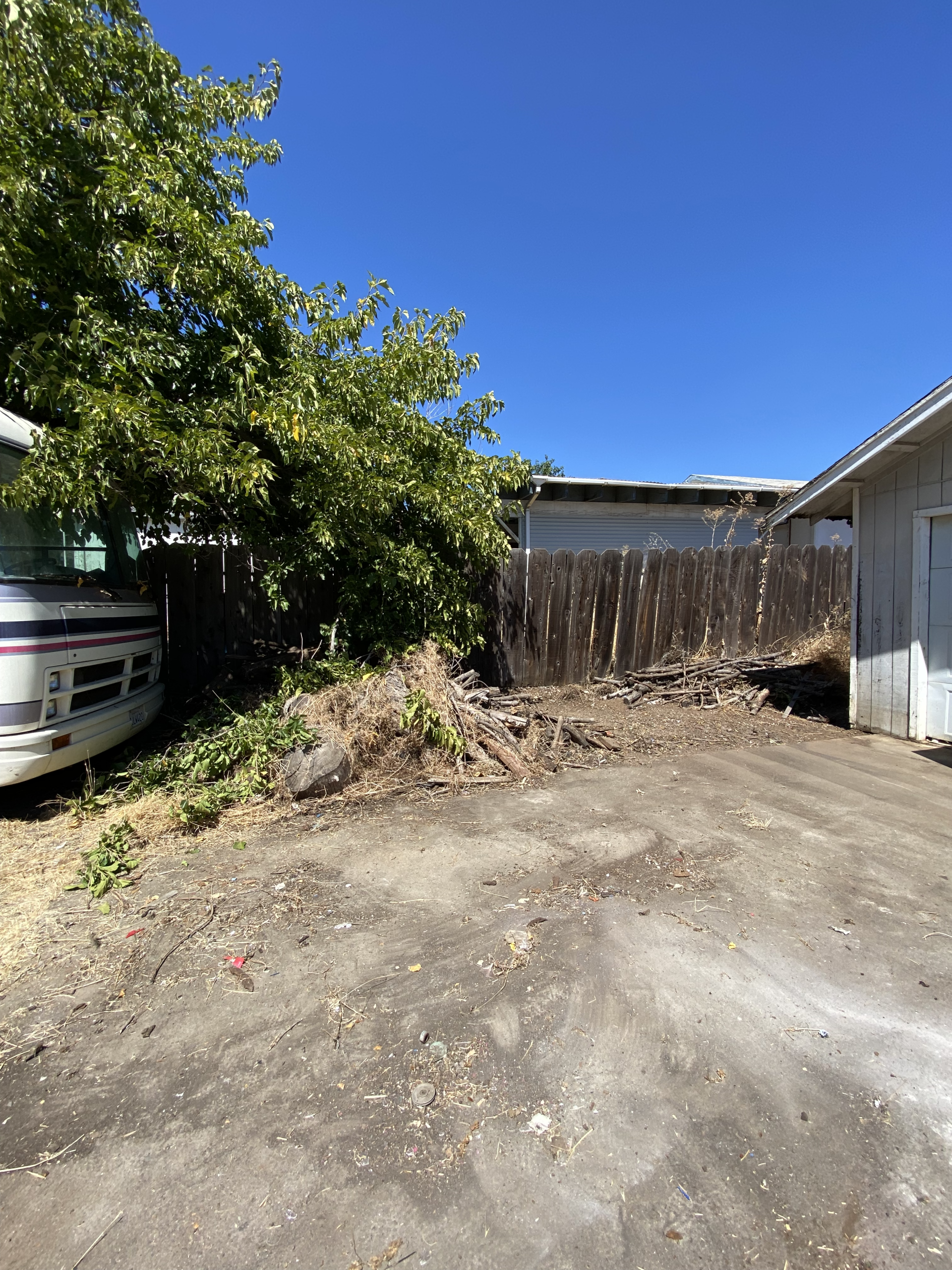 Dirt driveway beside a leafy tree, parked car, wooden fence, and white garage under a clear blue sky