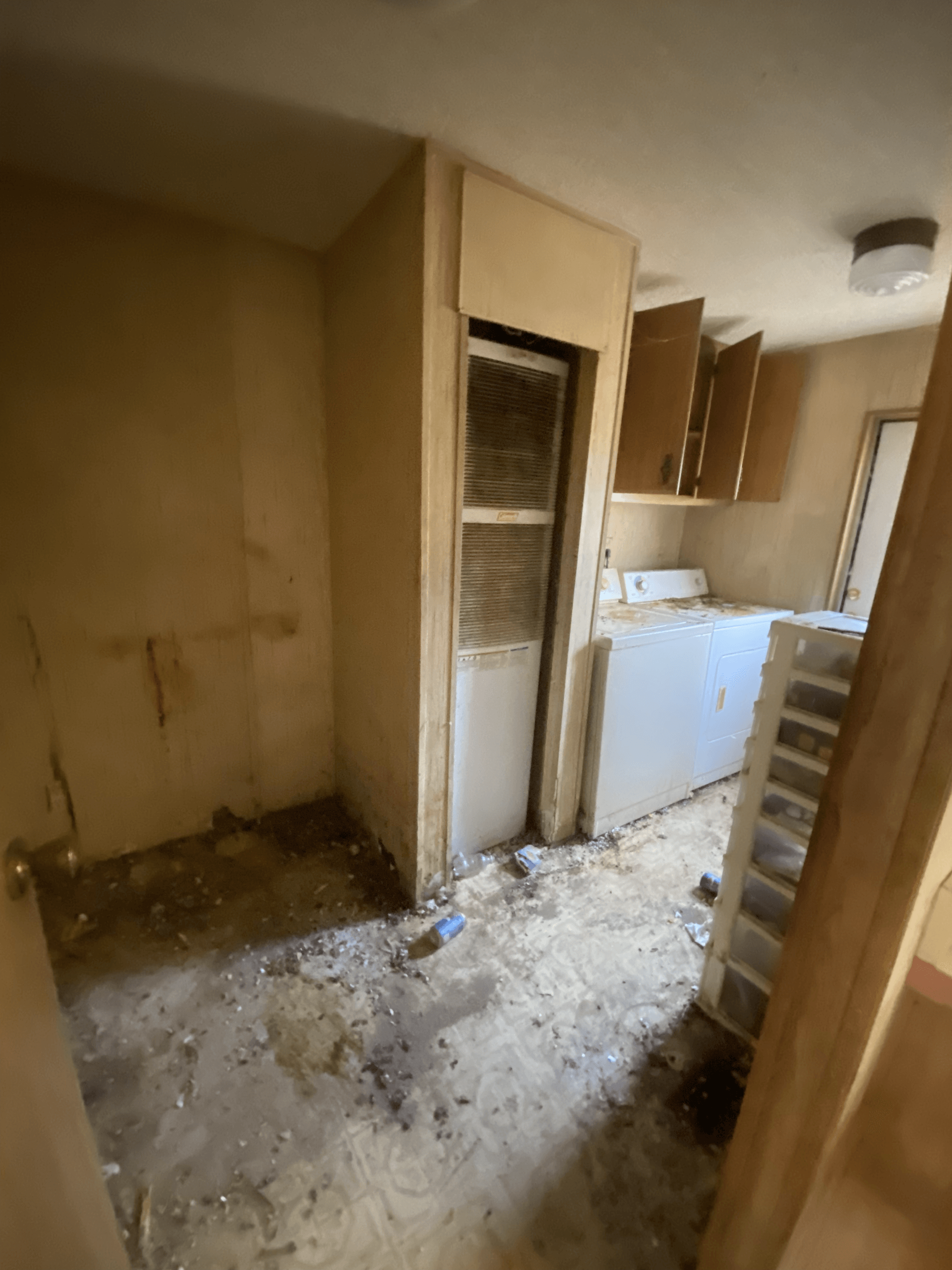 Damaged kitchen with exposed wall, debris-covered floor, and cabinets in a narrow hallway.