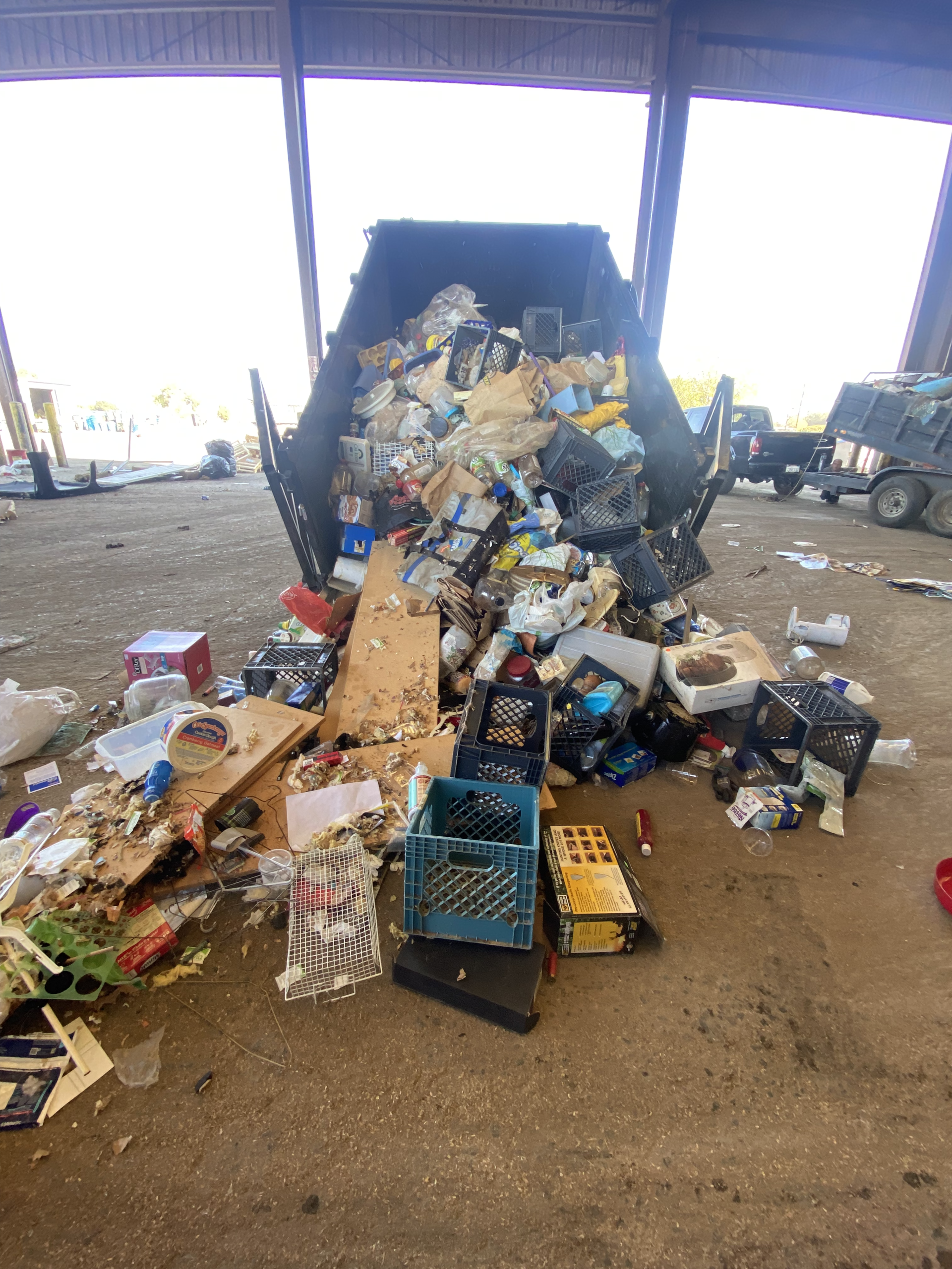 Overflowing dumpster at a covered waste facility, surrounded by scattered trash and recycling bins.