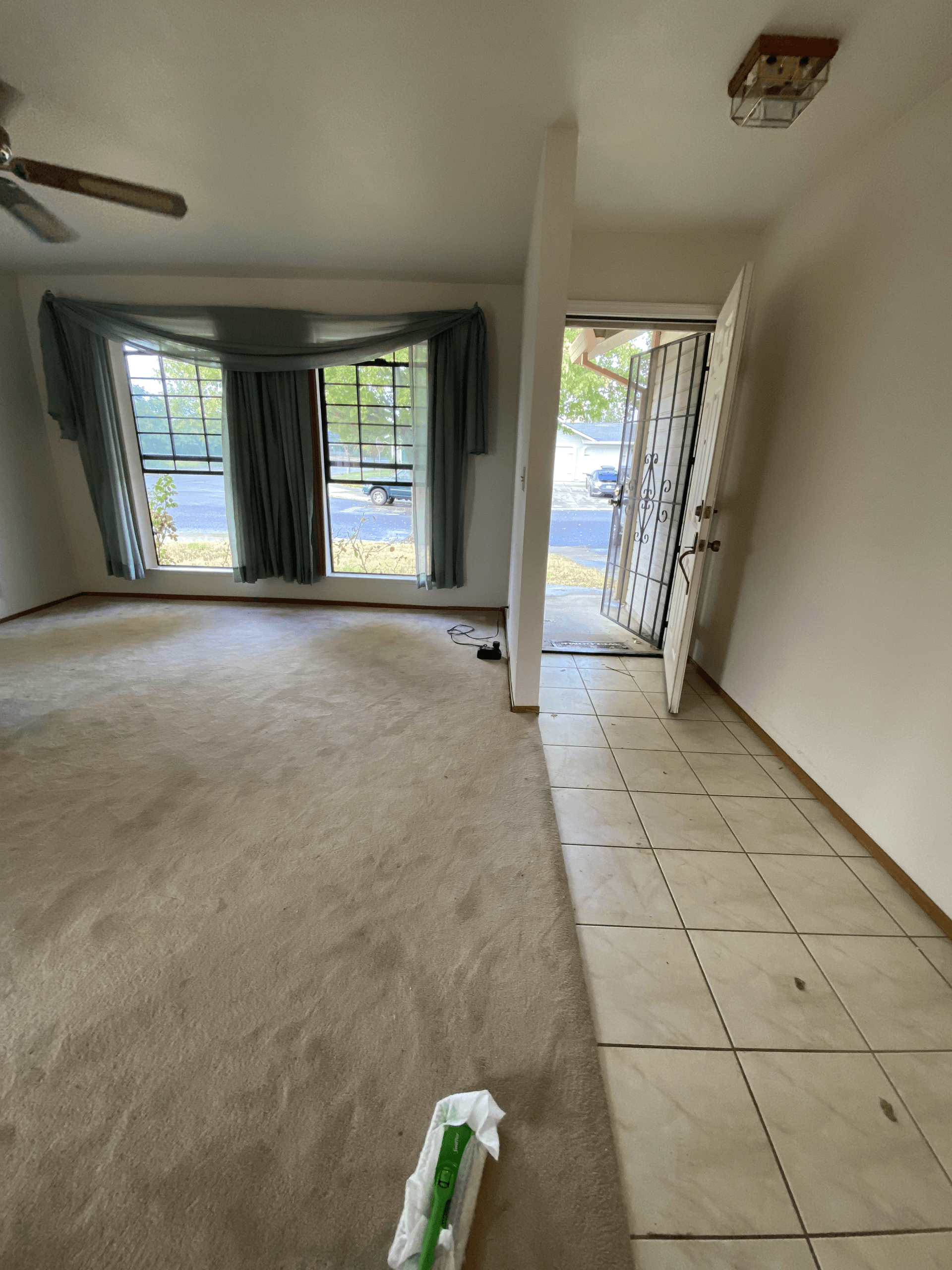 Empty living room with carpet, tiled entryway, curtained windows, and a ceiling fan.