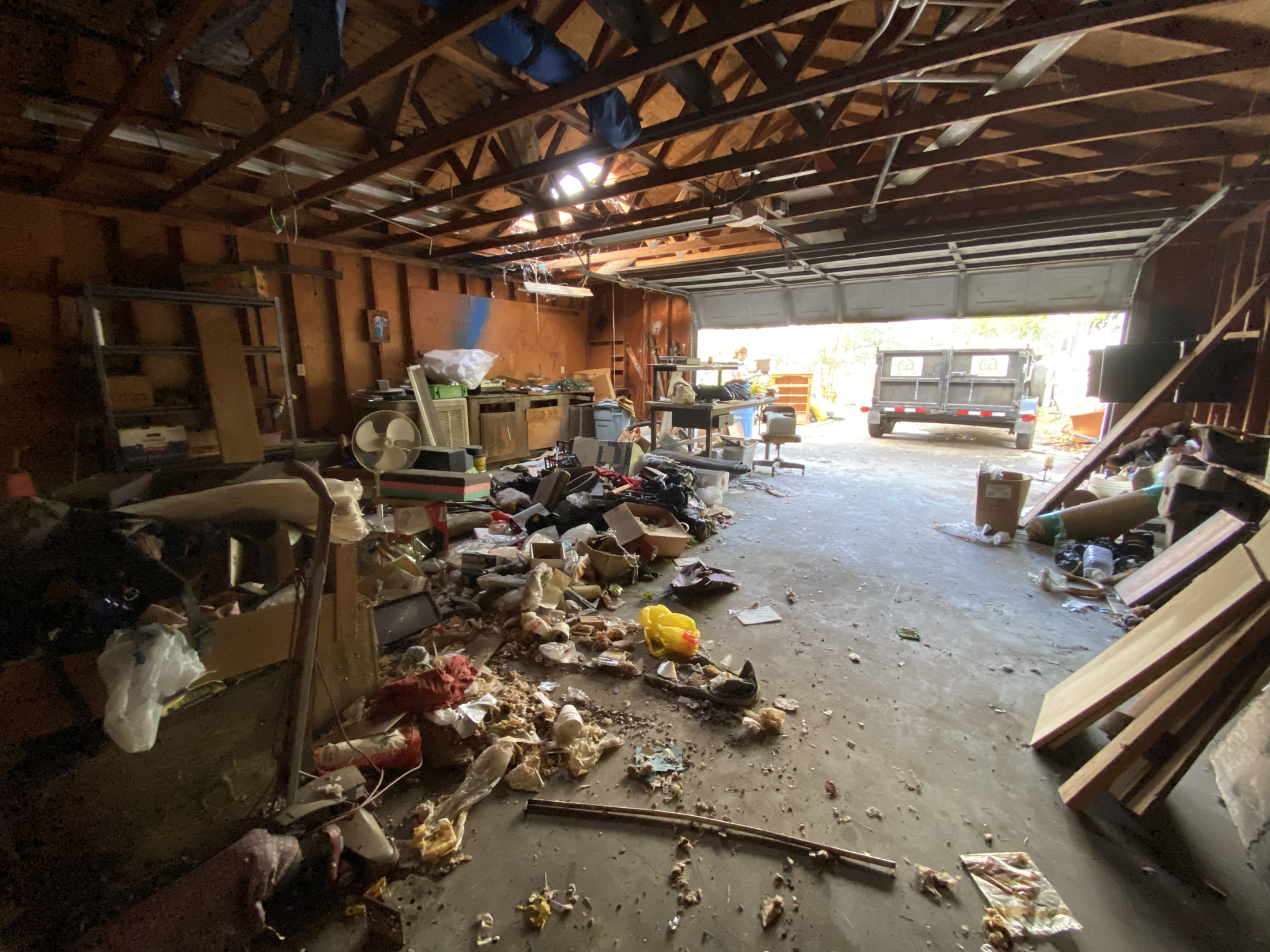 Cluttered garage with trash piles, tools, and a parked vehicle near the open doorway