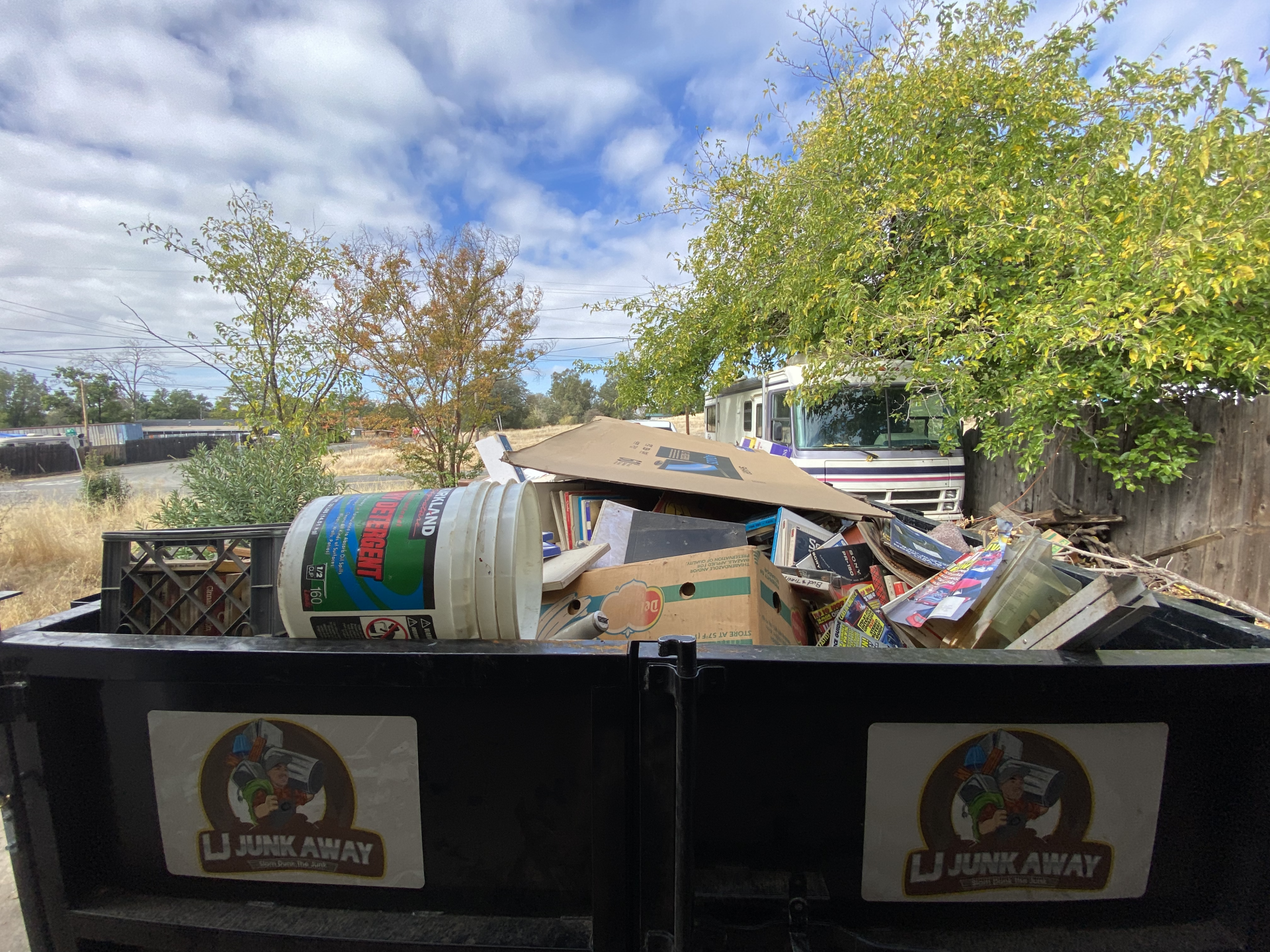 Overflowing dumpster with cardboard and debris outdoors beside a fence and trees