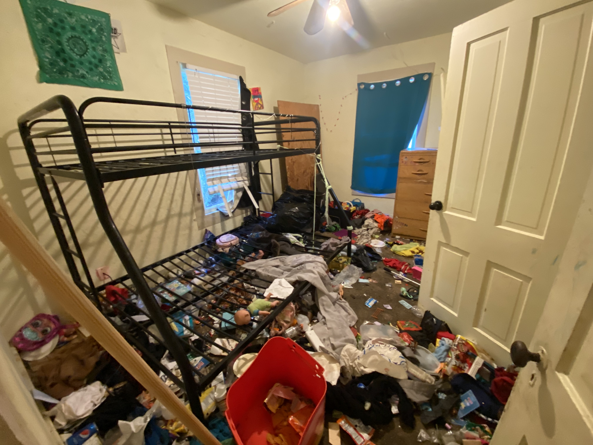 Cluttered bedroom with overturned metal bed frame, scattered clothes and debris, and a blue window curtain.