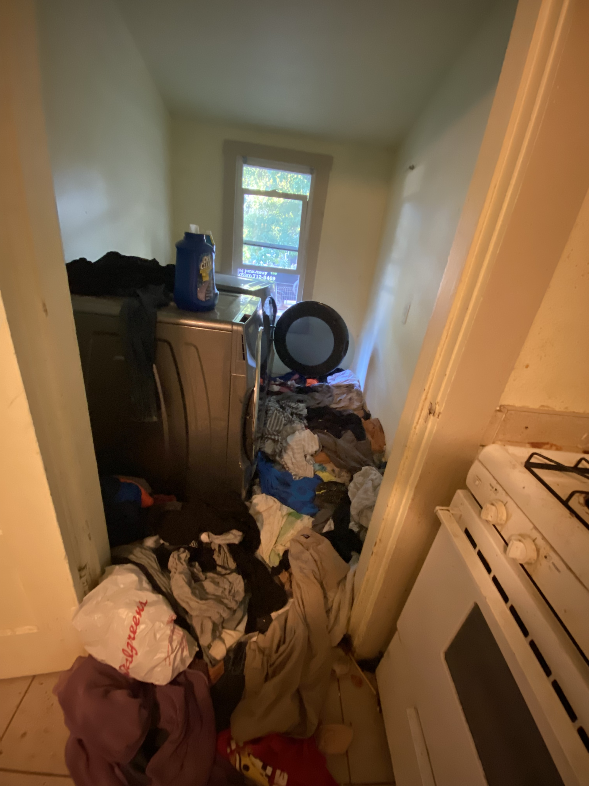 Cluttered narrow hallway with piles of clothes and belongings, a dresser, fan, and window at the end.