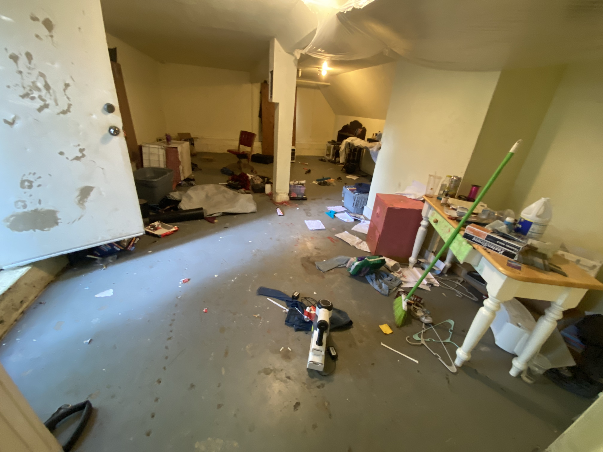 Cluttered room with scattered tools, debris, and furniture on a concrete floor; damaged, unfinished interior.