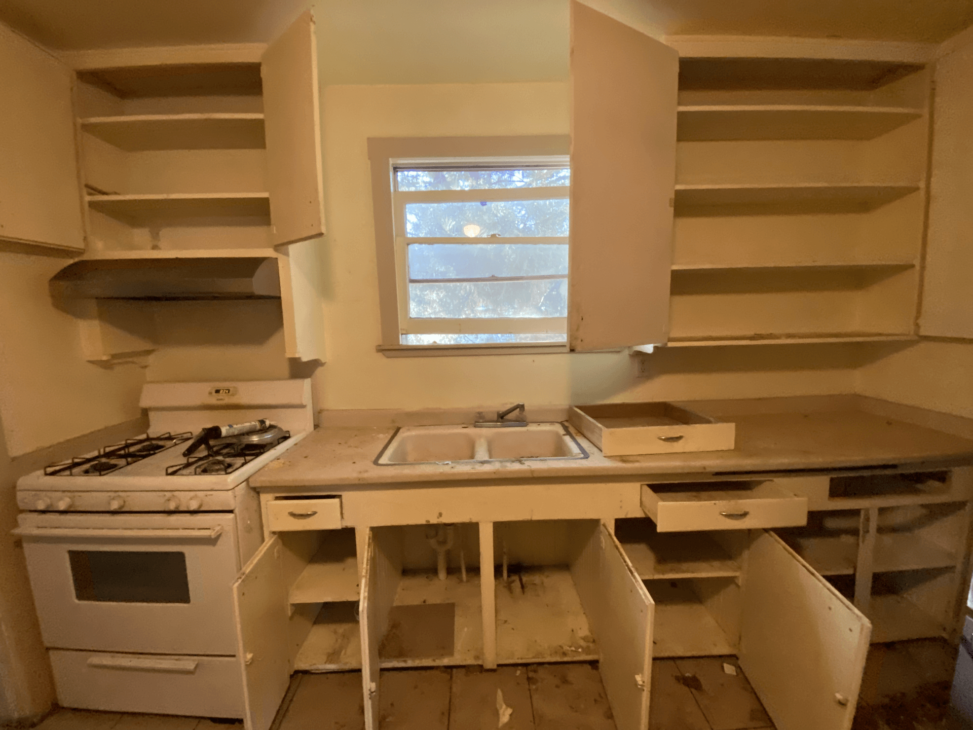 Empty beige kitchen with white cabinets, open shelves, a sink under a window, and a stove on the left.