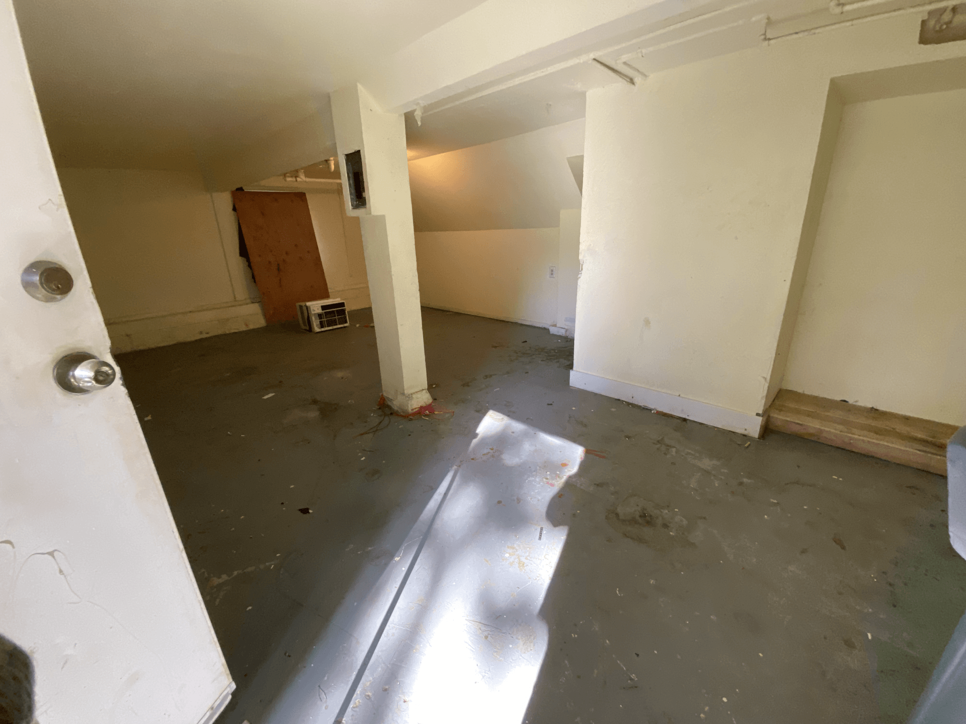 Empty unfinished room with beige walls, gray floor, and bright sunlight streaming through a window.