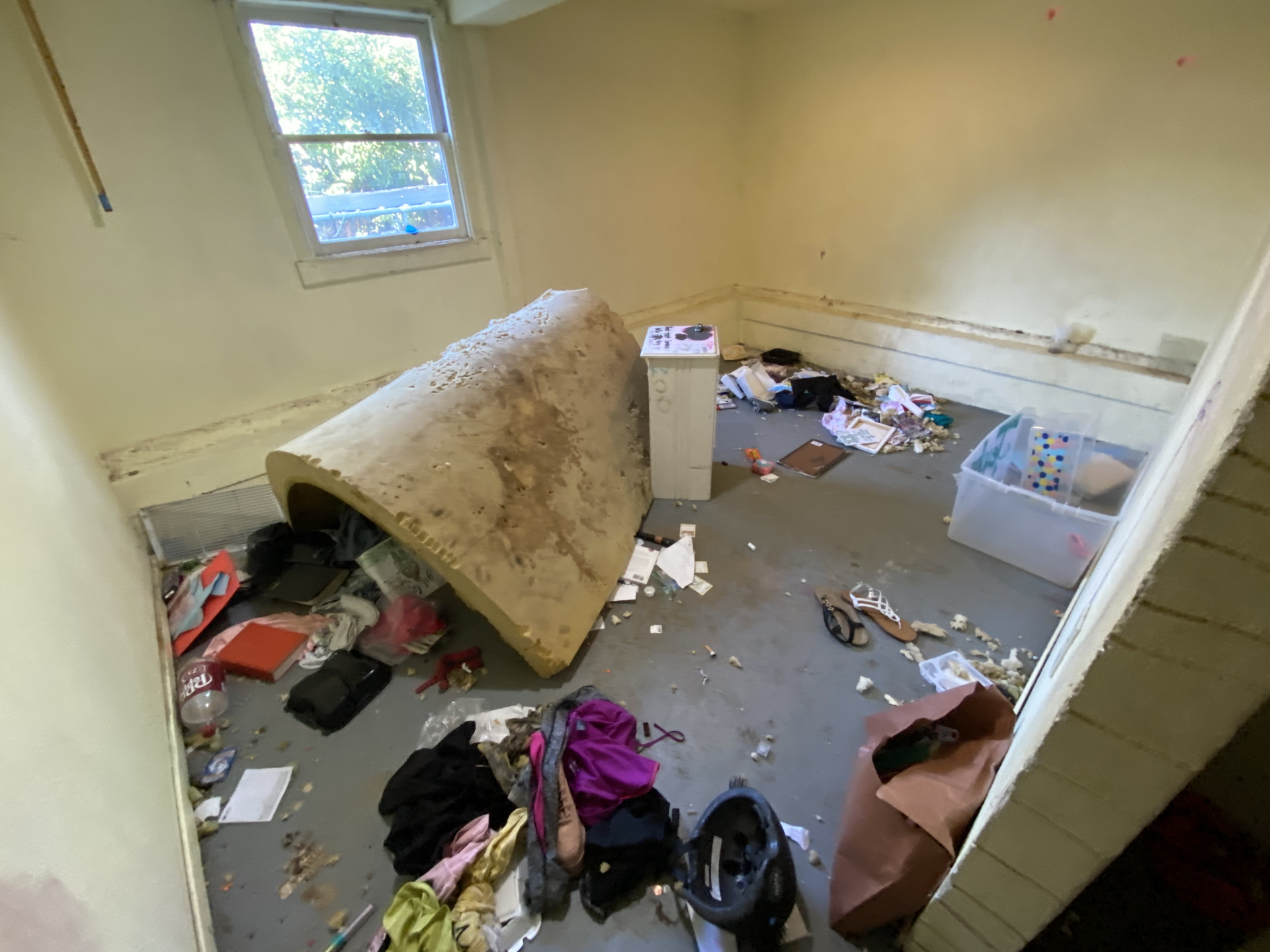 Cluttered, dilapidated room with debris, overturned furniture, and a rolled carpet under a window.