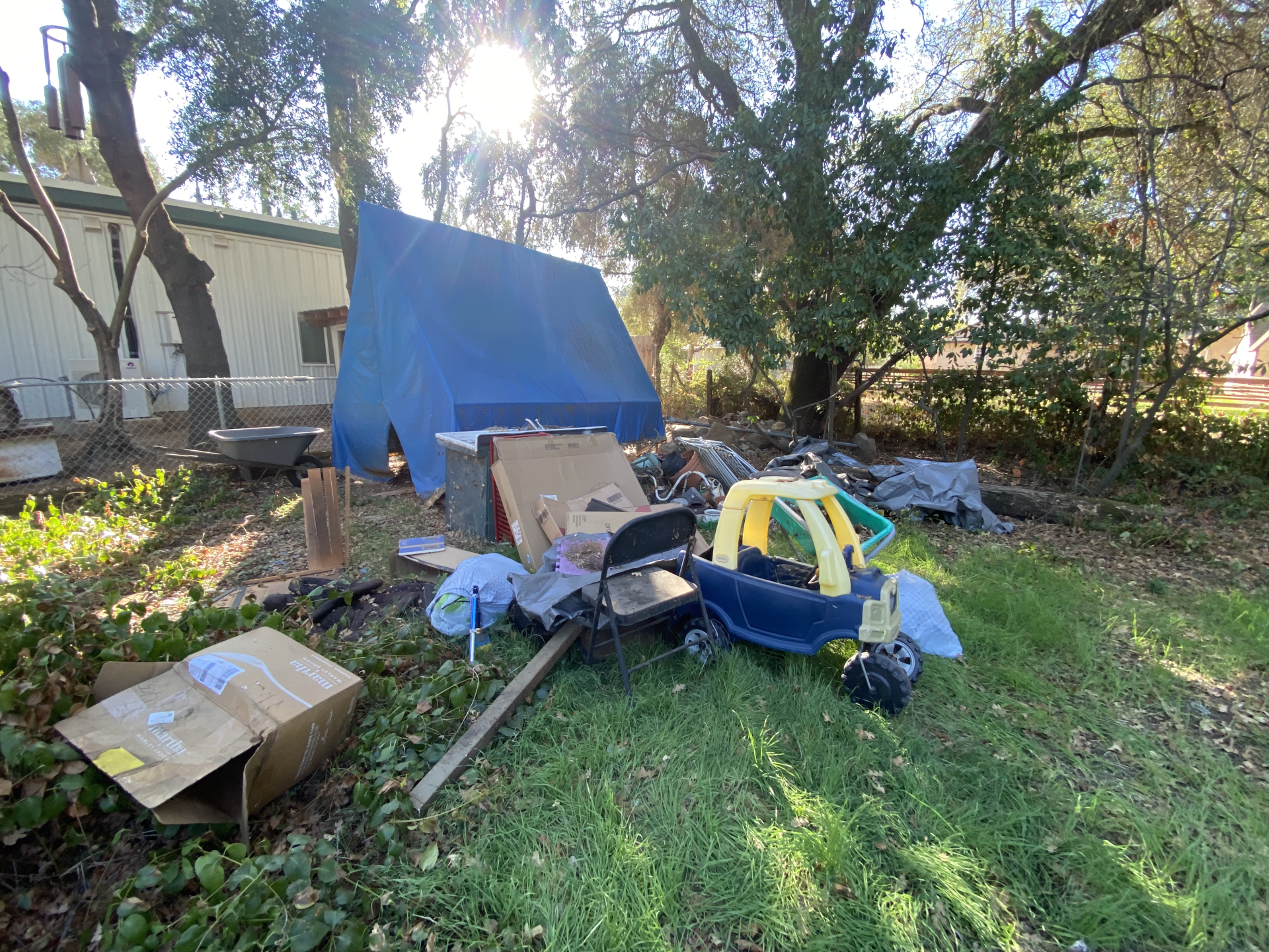 Cluttered backyard with tarp, boxes, and scattered tools on grass in sunlight