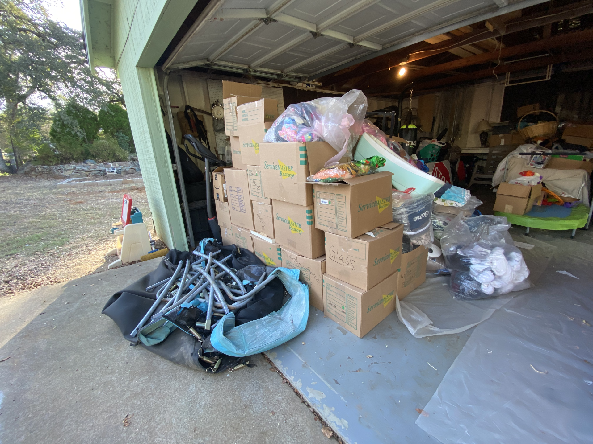 Cluttered garage with stacked cardboard boxes, plastic bags, and assorted household items near the open door