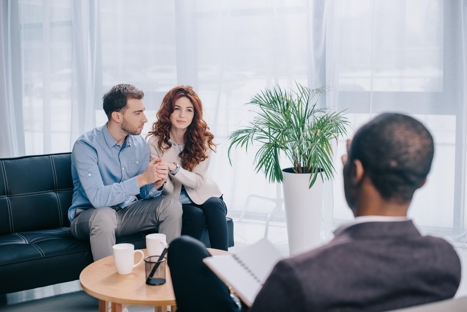 Couple sitting on couch, holding hands, talking to a therapist in an office setting.